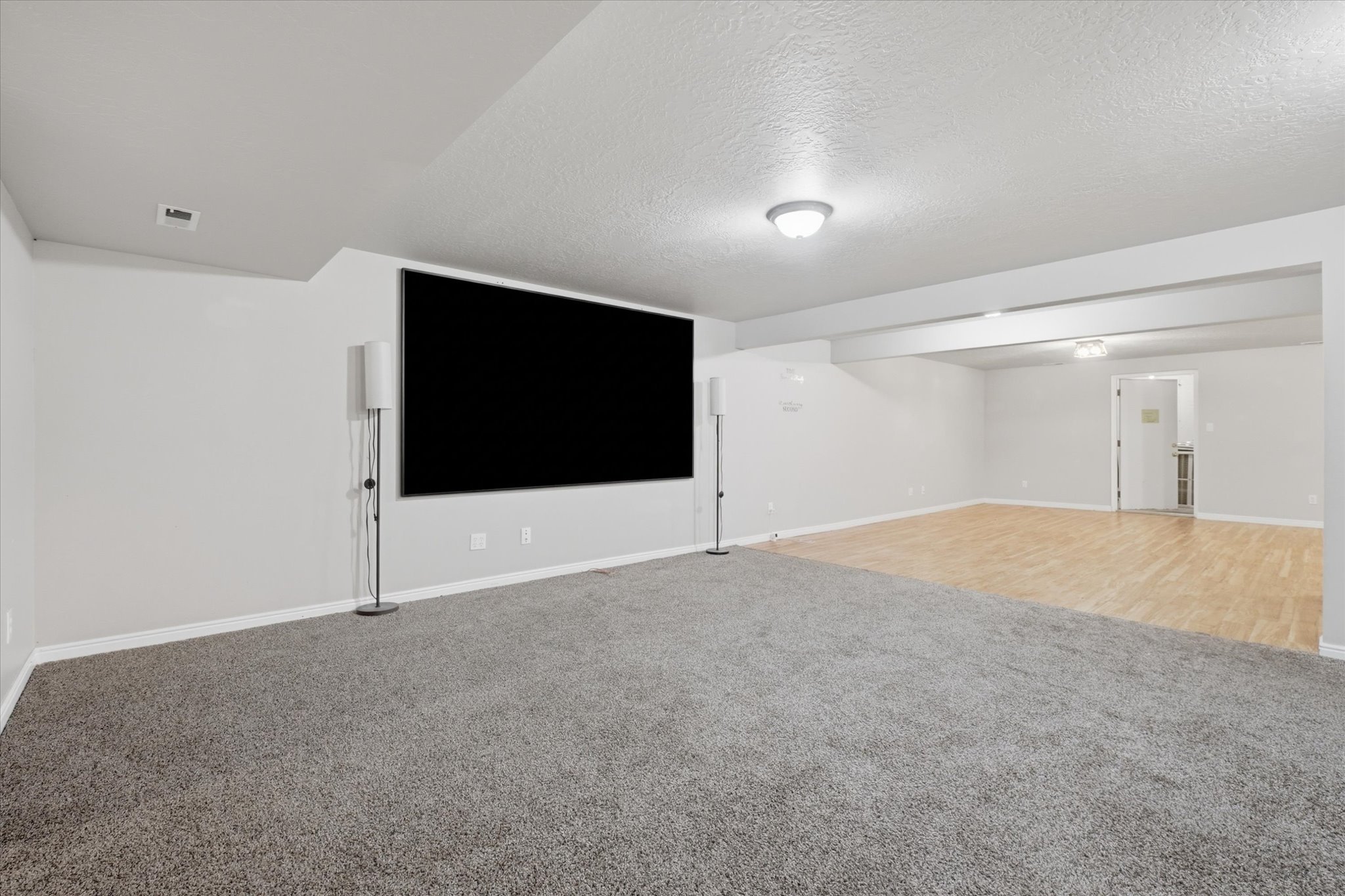 Open recreational space featuring a large projection screen, neutral gray carpeting, and light wood-finish flooring