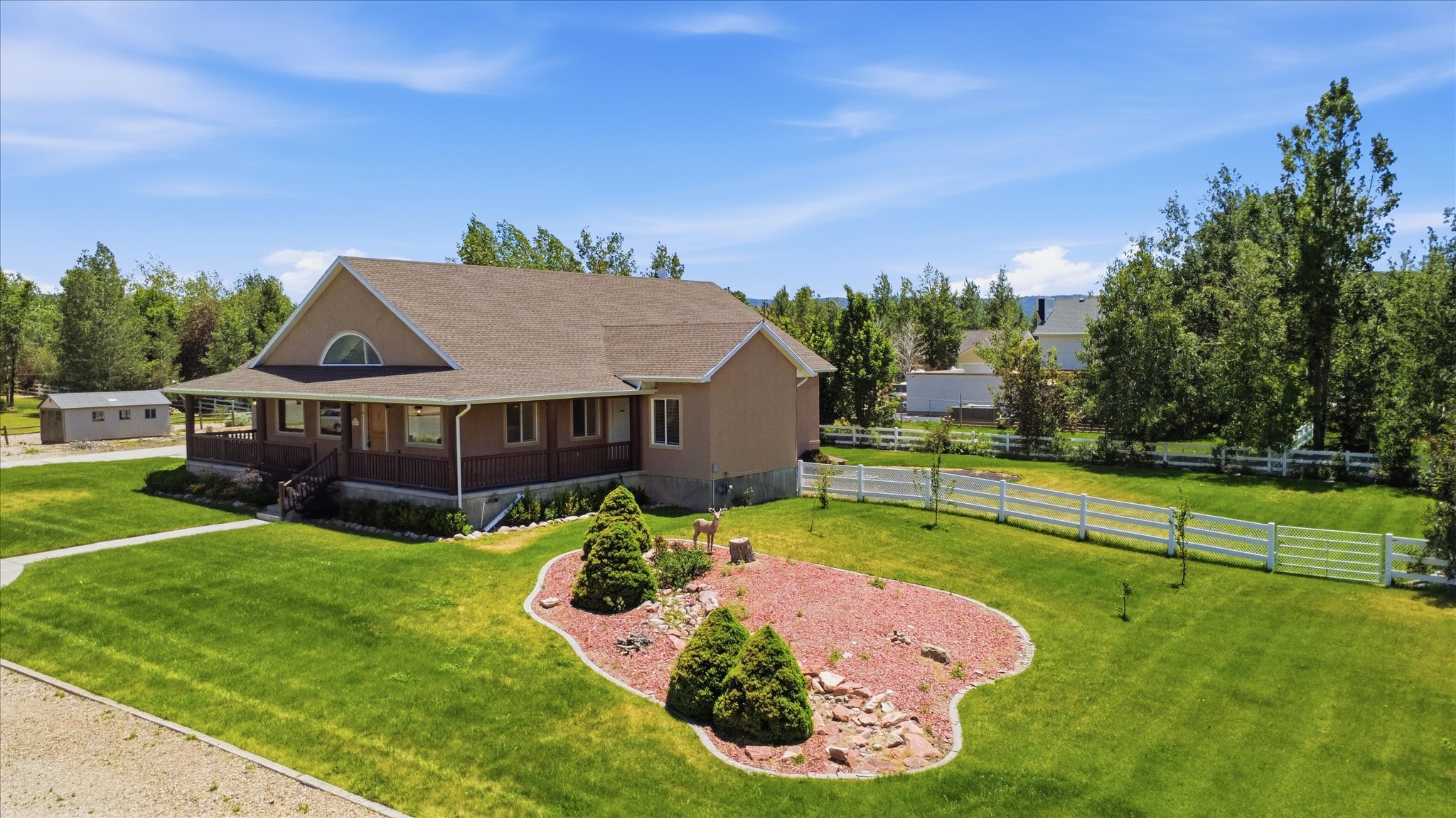 Expansive property featuring a tan exterior, a broad covered porch, a manicured lawn, a decorative landscaped bed, and a white picket fence