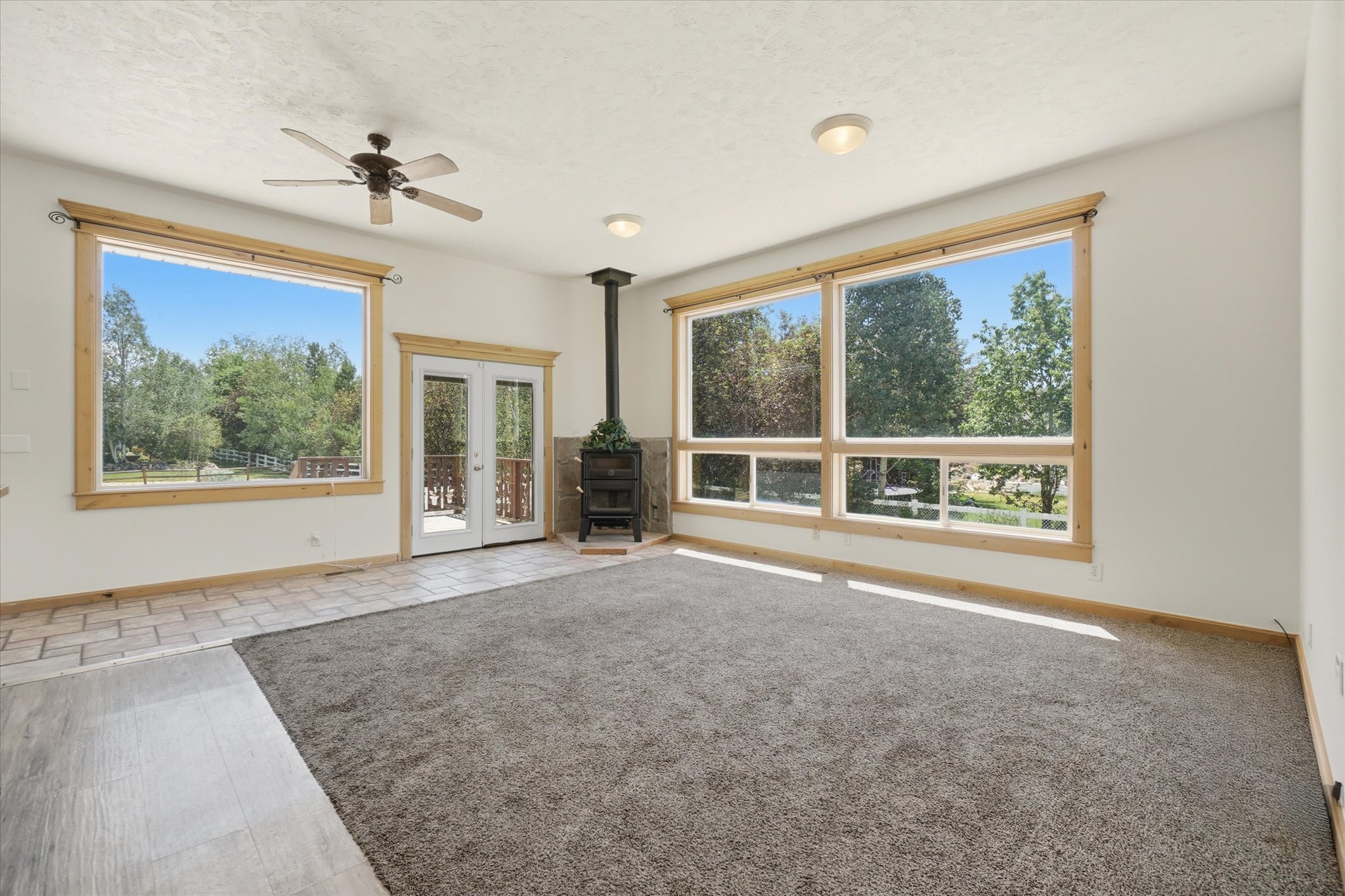 Spacious living area featuring large windows with natural wood trim, a central wood-burning stove, and a ceiling fan