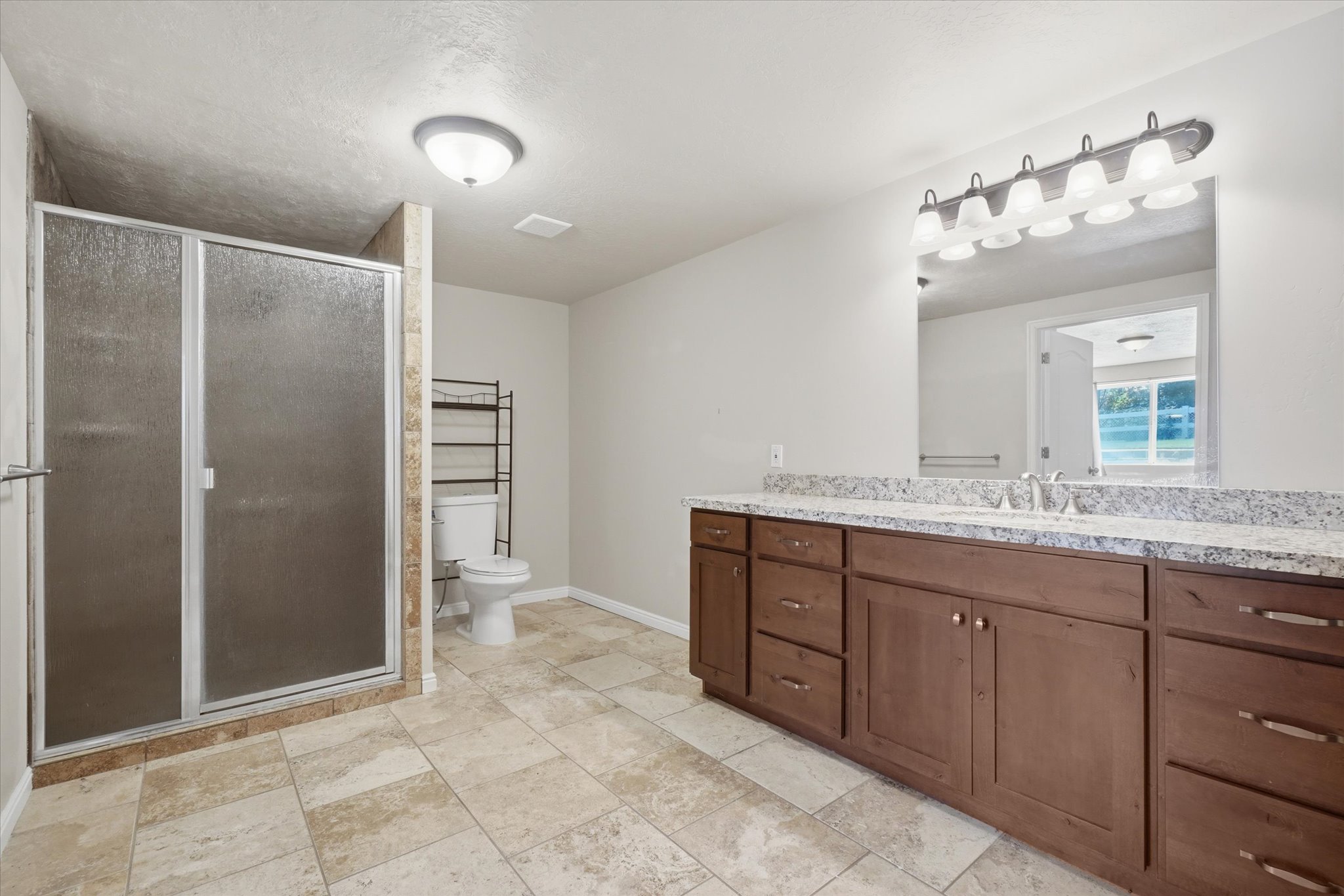 Bathroom featuring a spacious walk-in shower with frosted glass doors, a large vanity with a double sink and ample storage, stone-look tile flooring, and a modern light fixture above the vanity
