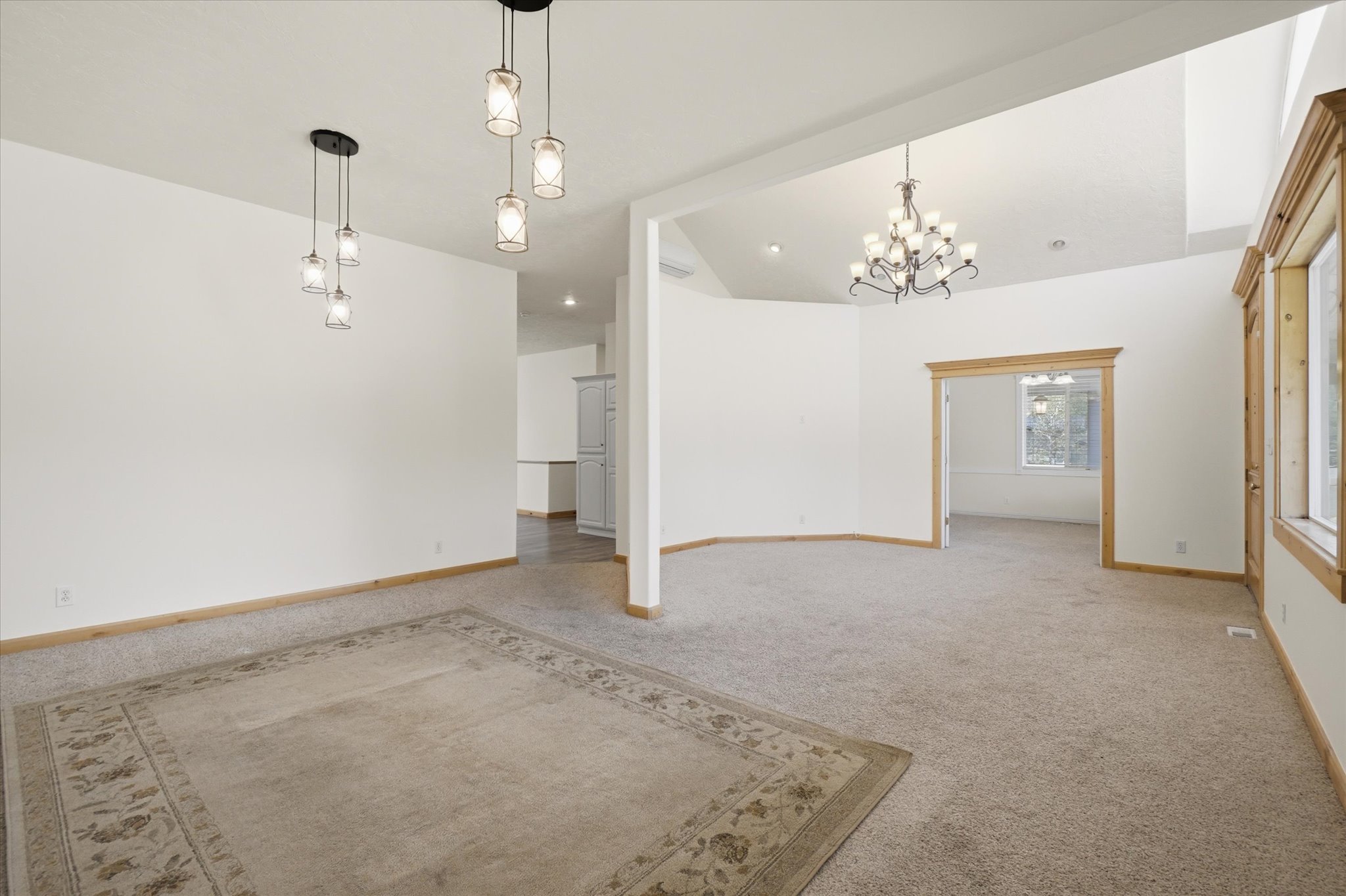 Spacious carpeted interior featuring multiple pendant light fixtures, a chandelier, wood trim, and a vaulted ceiling