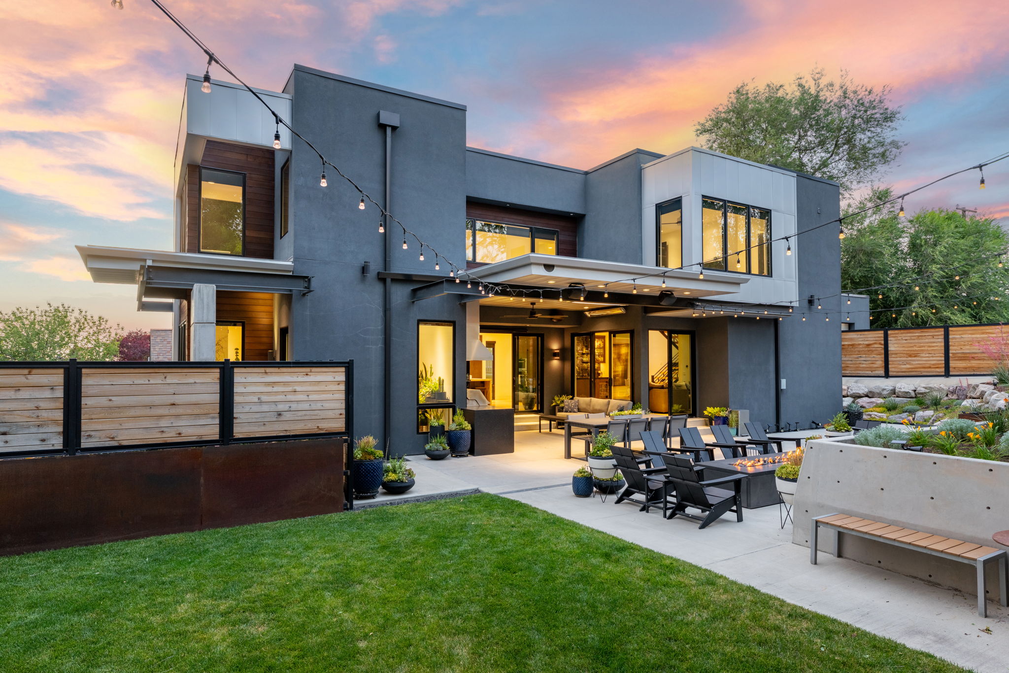 Modern architectural exterior featuring multiple large windows, a spacious concrete patio, integrated fire pit, and a manicured lawn