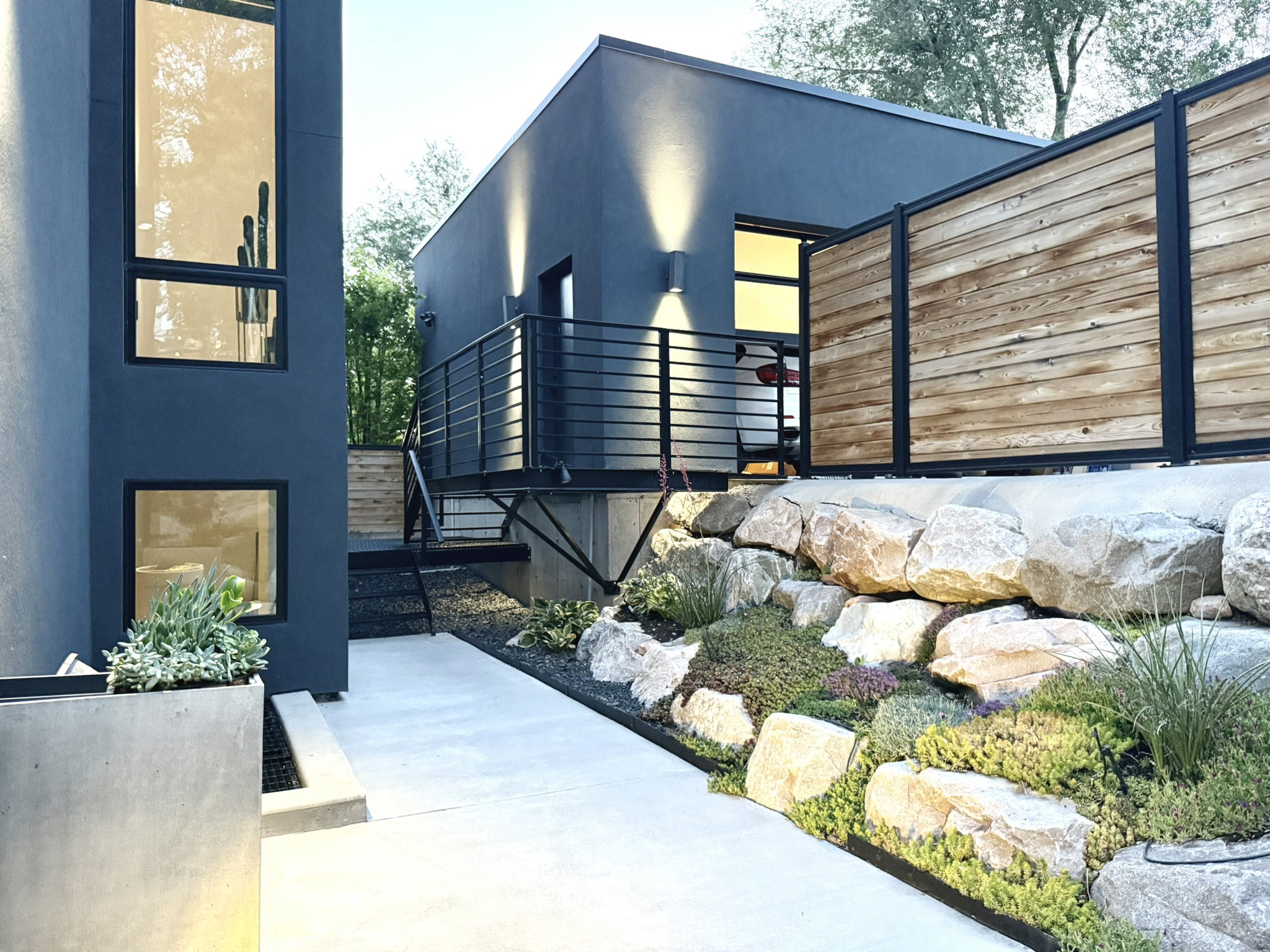 Modern exterior featuring dark gray stucco, a concrete walkway, a tiered rock garden with plantings, a wood and metal privacy fence, and an elevated patio with metal railings and sconce lighting