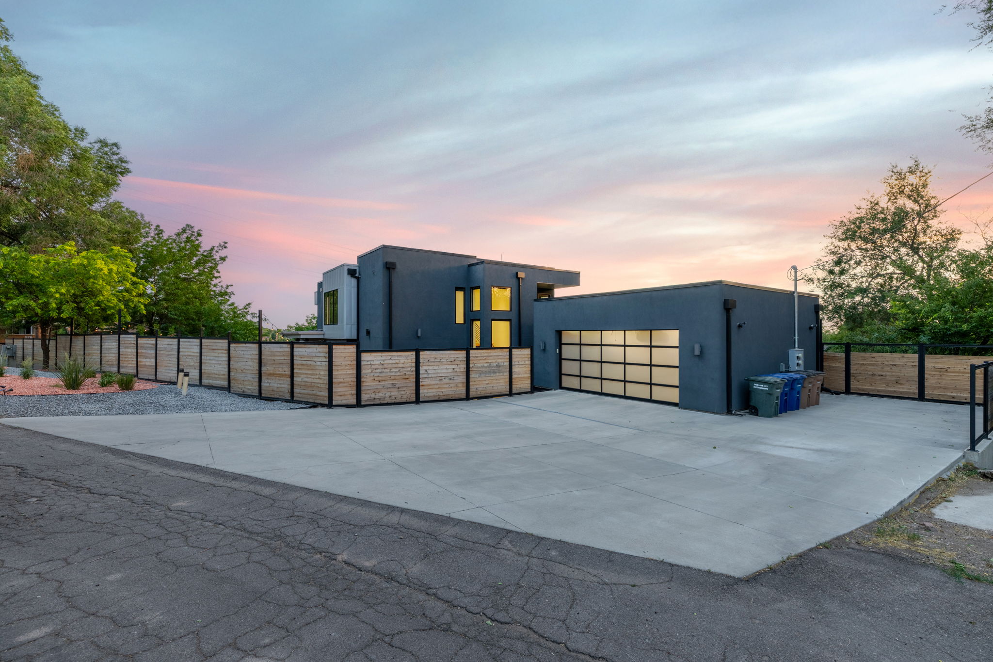 Contemporary exterior with dark stucco finish, a full-view garage door, a private wood-panel fence, and a large concrete driveway