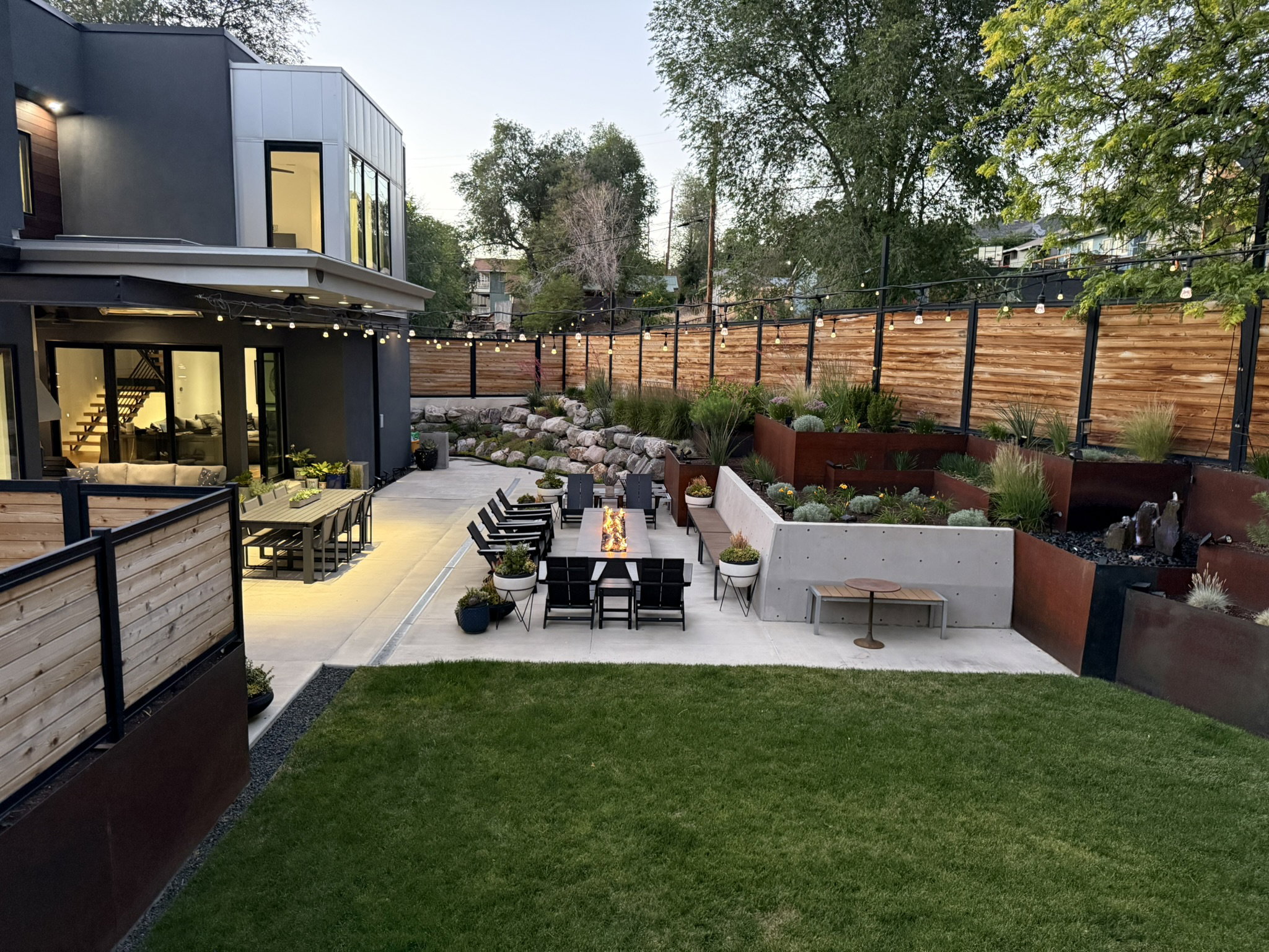Expansive hardscaped patio featuring multiple seating areas, a built-in fire feature, and decorative string lighting