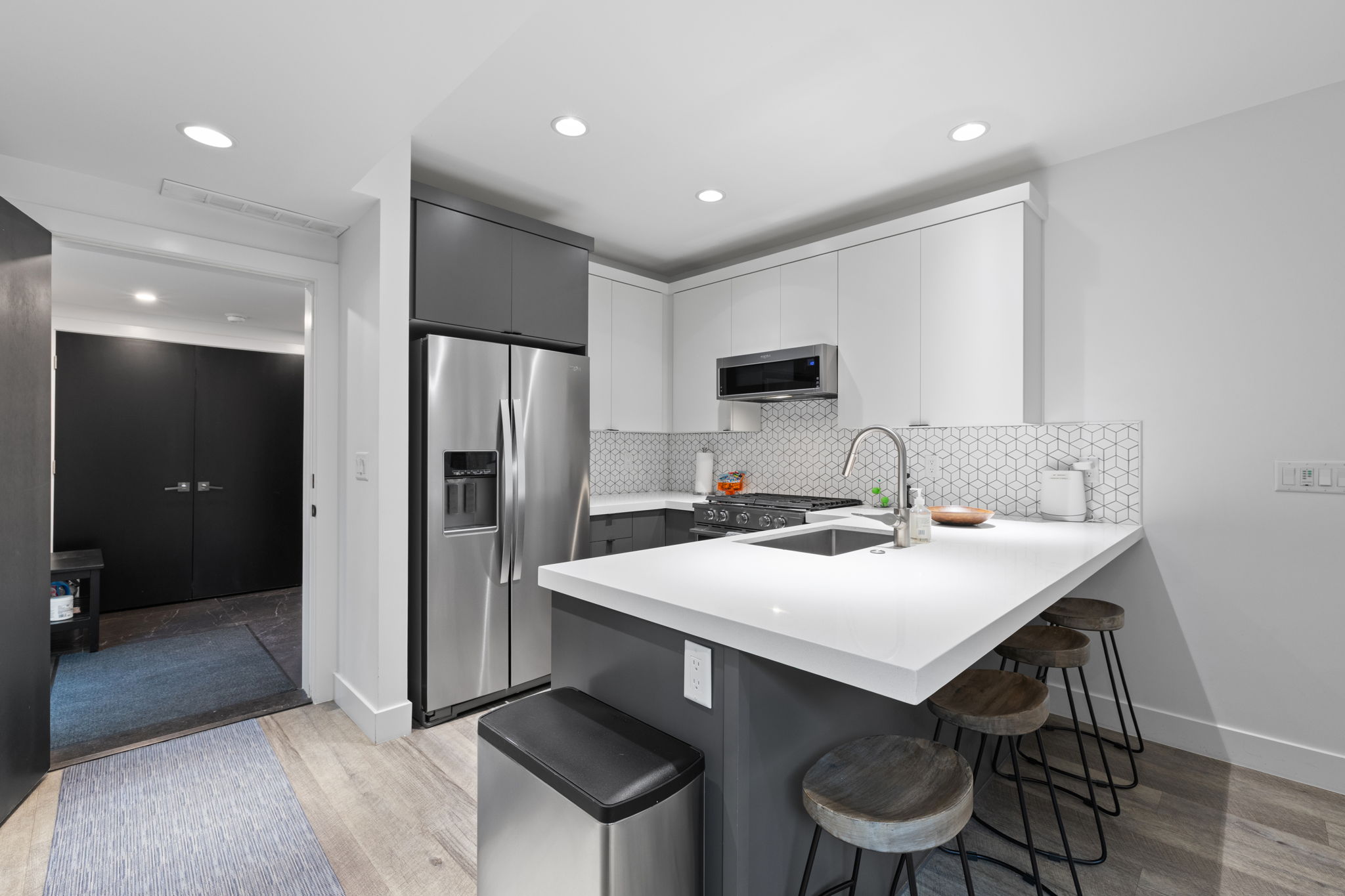 Contemporary kitchen featuring a white countertop island with integrated sink, stainless steel appliances, two-tone cabinetry, and geometric tile backsplash