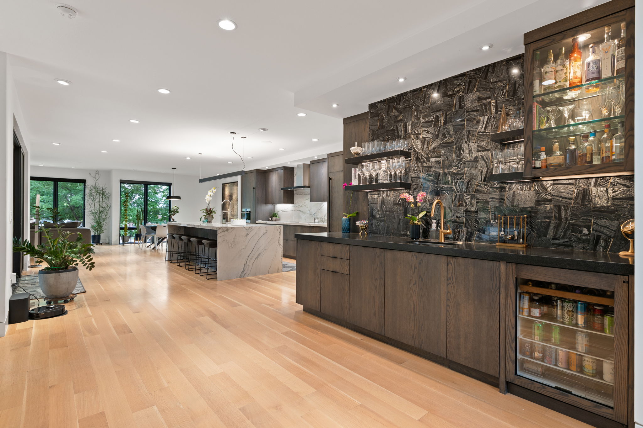 Expansive entertaining space featuring light wood-finish flooring, a full wet bar with dark stone backsplash and under-counter beverage refrigeration, and a waterfall island with counter seating