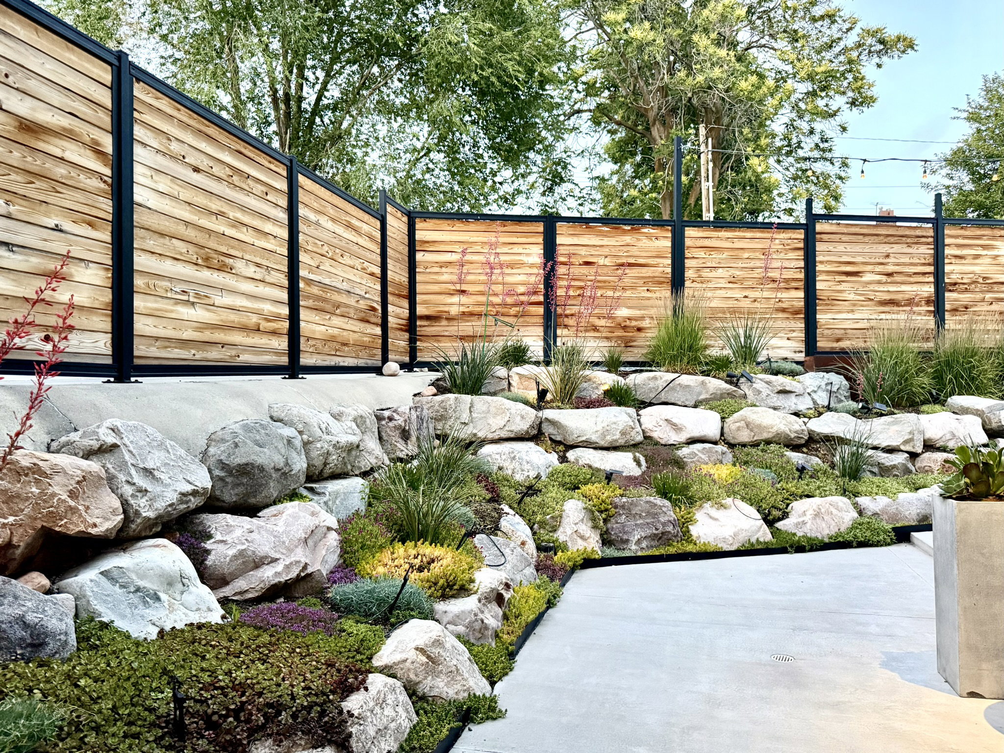 Outdoor hardscaping featuring a concrete patio, a tiered rock garden with diverse plantings, and a horizontal wood-slat fence with dark metal posts