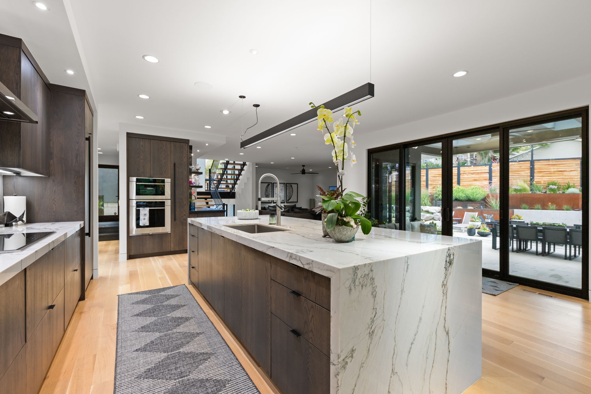 Modern kitchen featuring a waterfall island with a rectangular undermount sink, light wood-finish flooring, dark wood-finish cabinetry, built-in stainless steel appliances, and large sliding glass doors opening to an outdoor patio