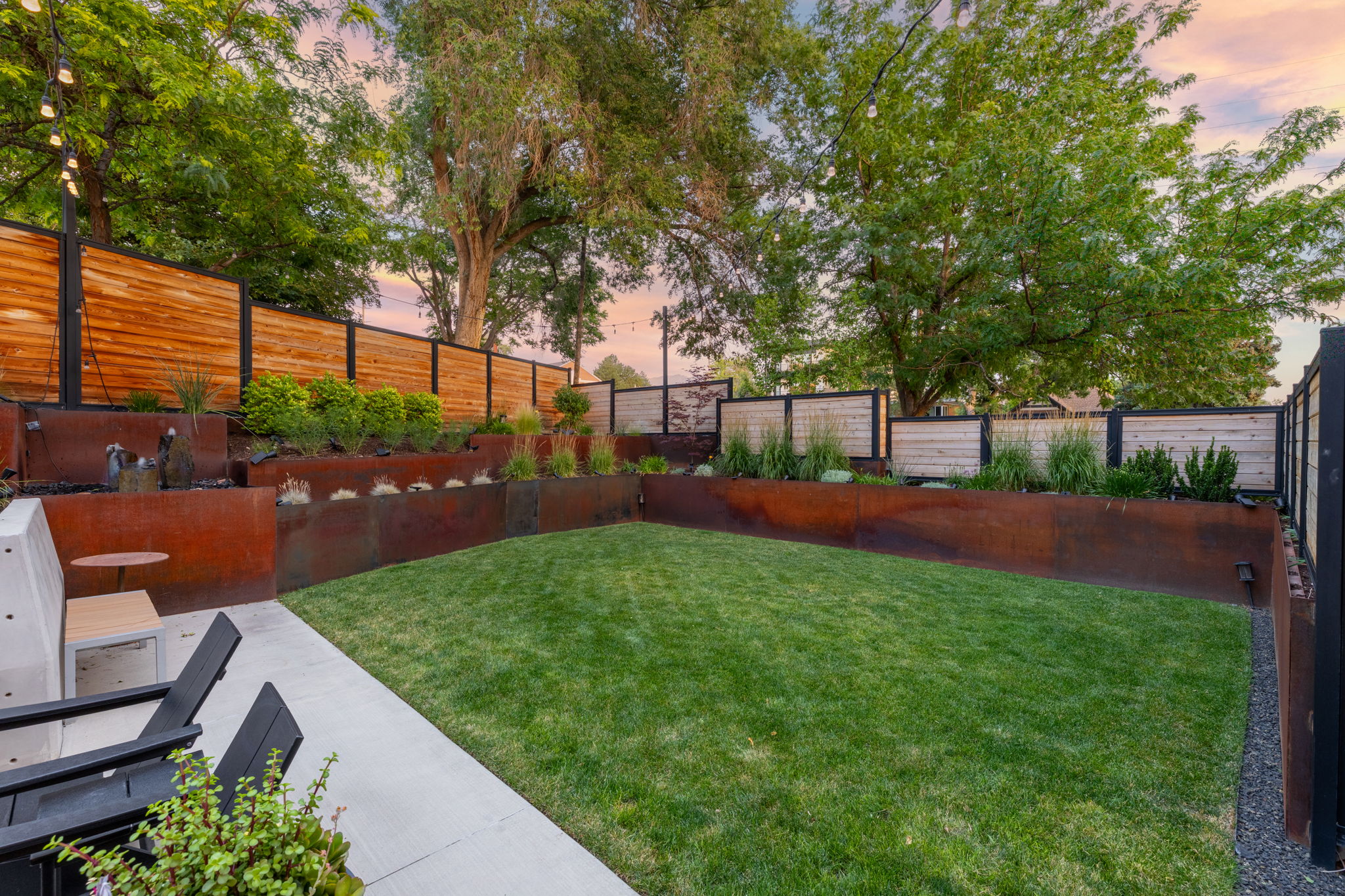 Expansive backyard featuring a lush lawn, concrete patio, multi-tiered corten steel retaining walls, layered landscaping, and horizontal wood fencing with black metal posts
