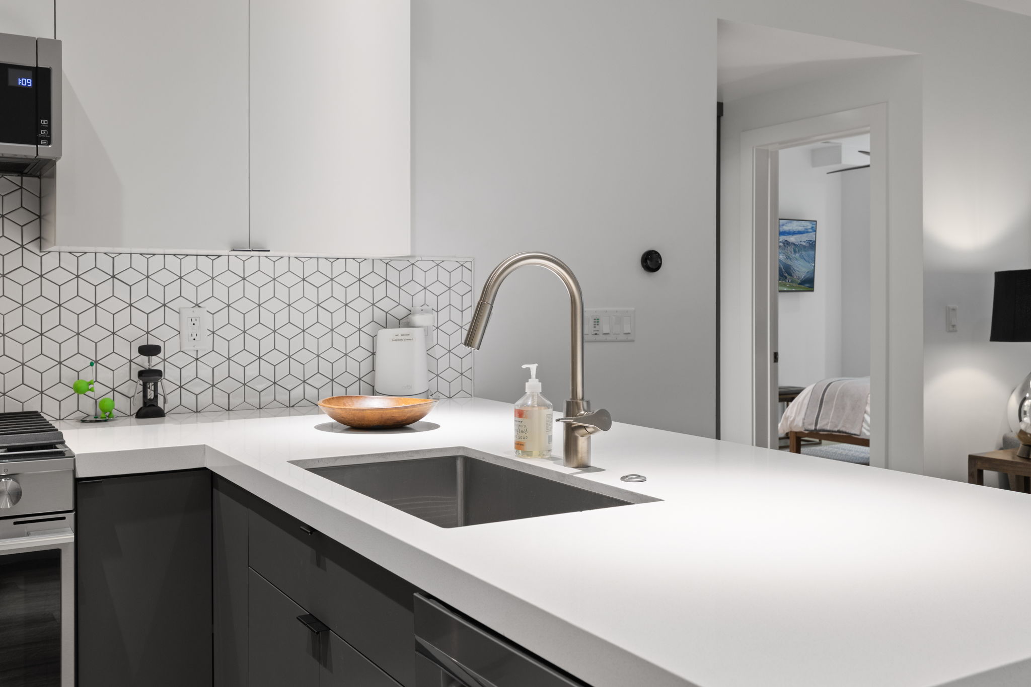 Modern kitchen featuring white countertops, stainless steel sink with gooseneck faucet, geometric pattern tile backsplash, white upper cabinetry, and dark lower cabinetry