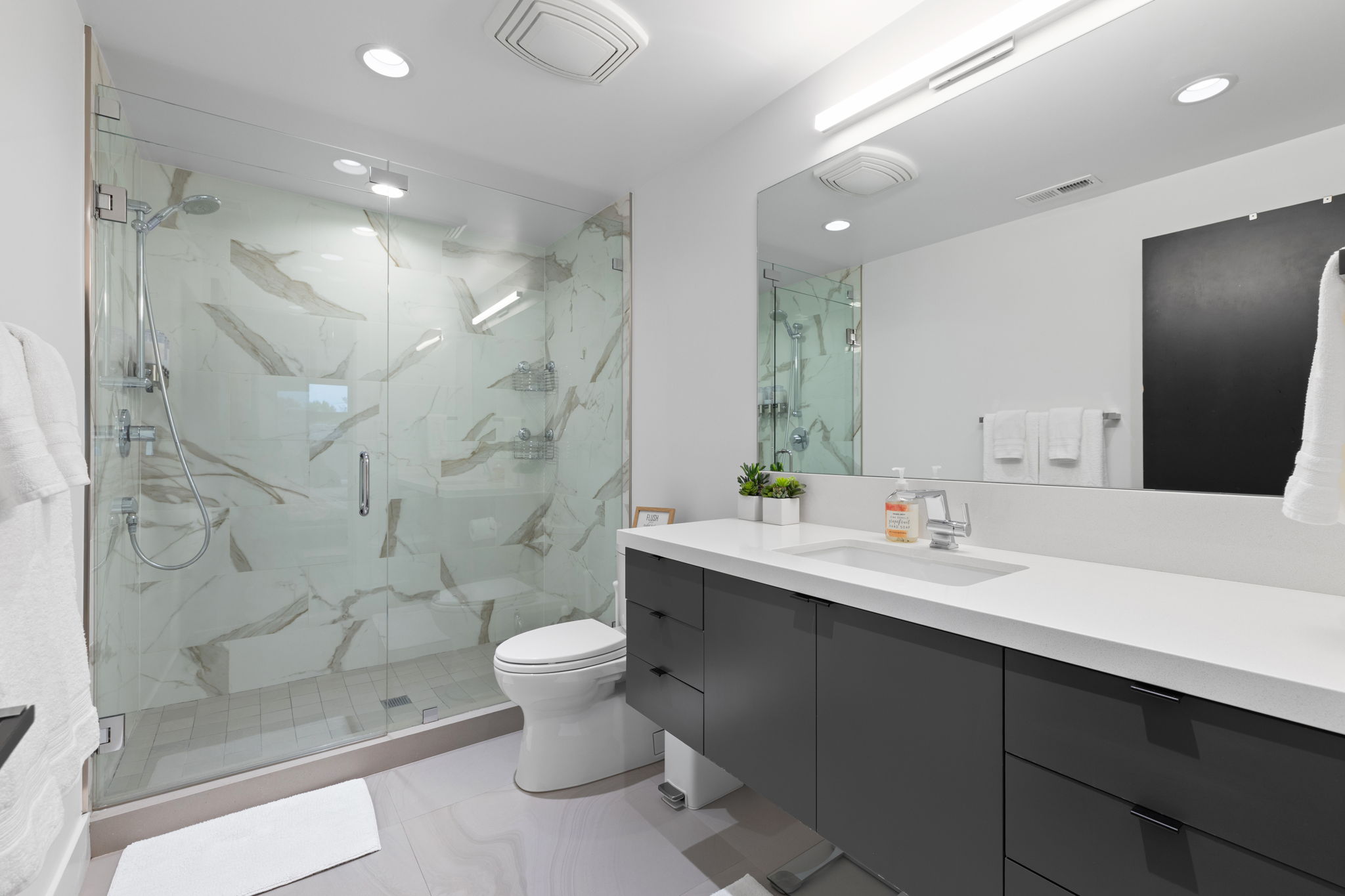 Spacious bathroom featuring a glass-enclosed shower with large format tile, a dual vanity with a white countertop, and contemporary cabinetry