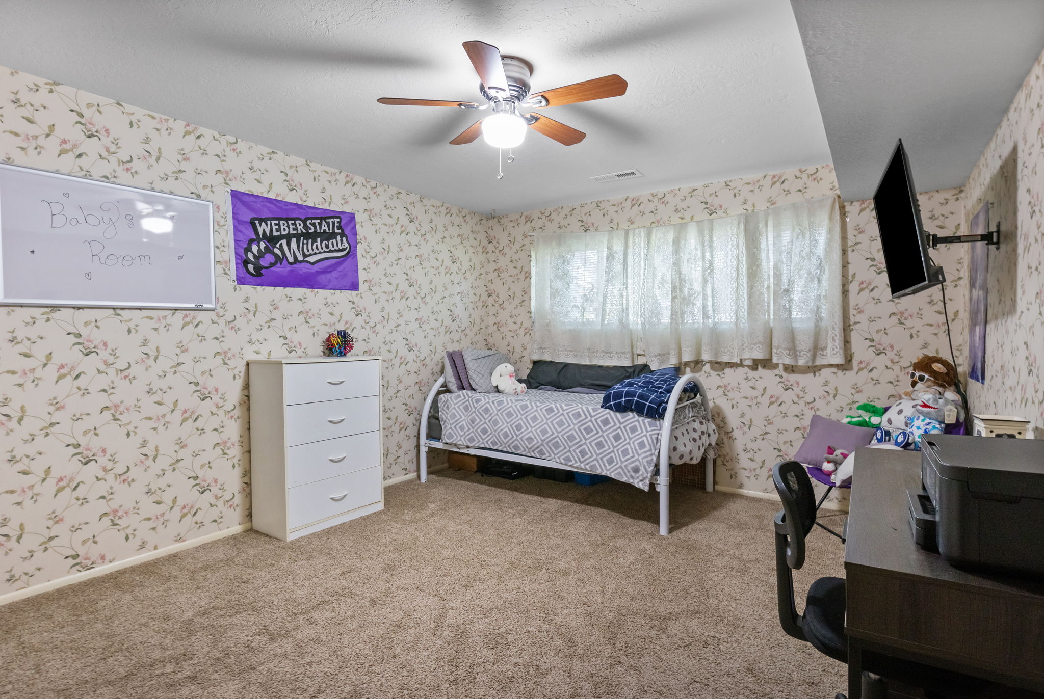 Carpeted room featuring patterned wallpaper, a ceiling fan with light fixture, and window with sheer curtains