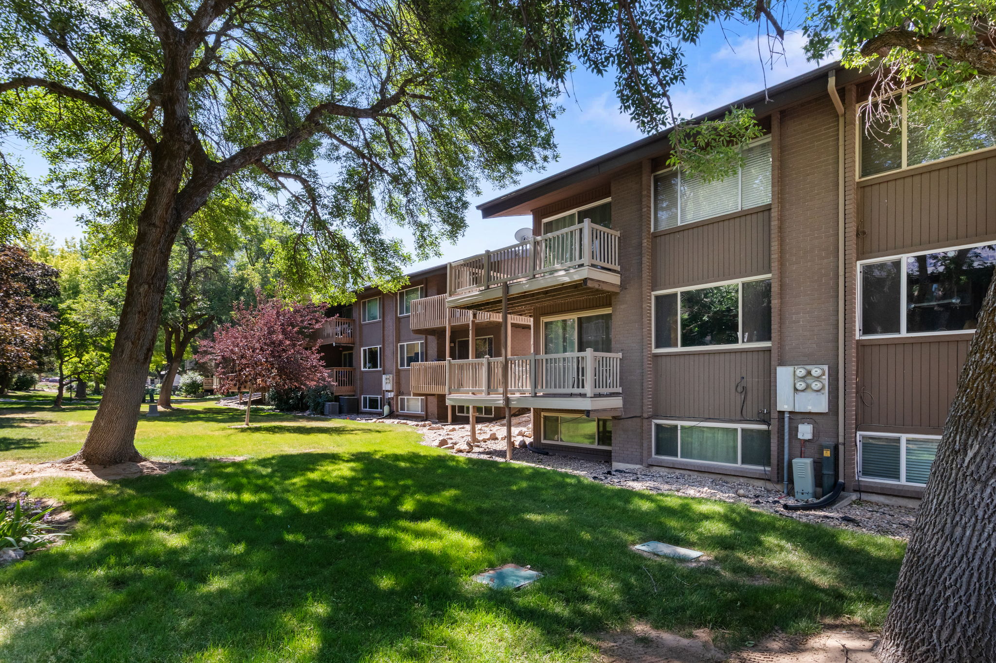 Multi-story building featuring private balconies, brick accents, and a surrounding green space with mature trees