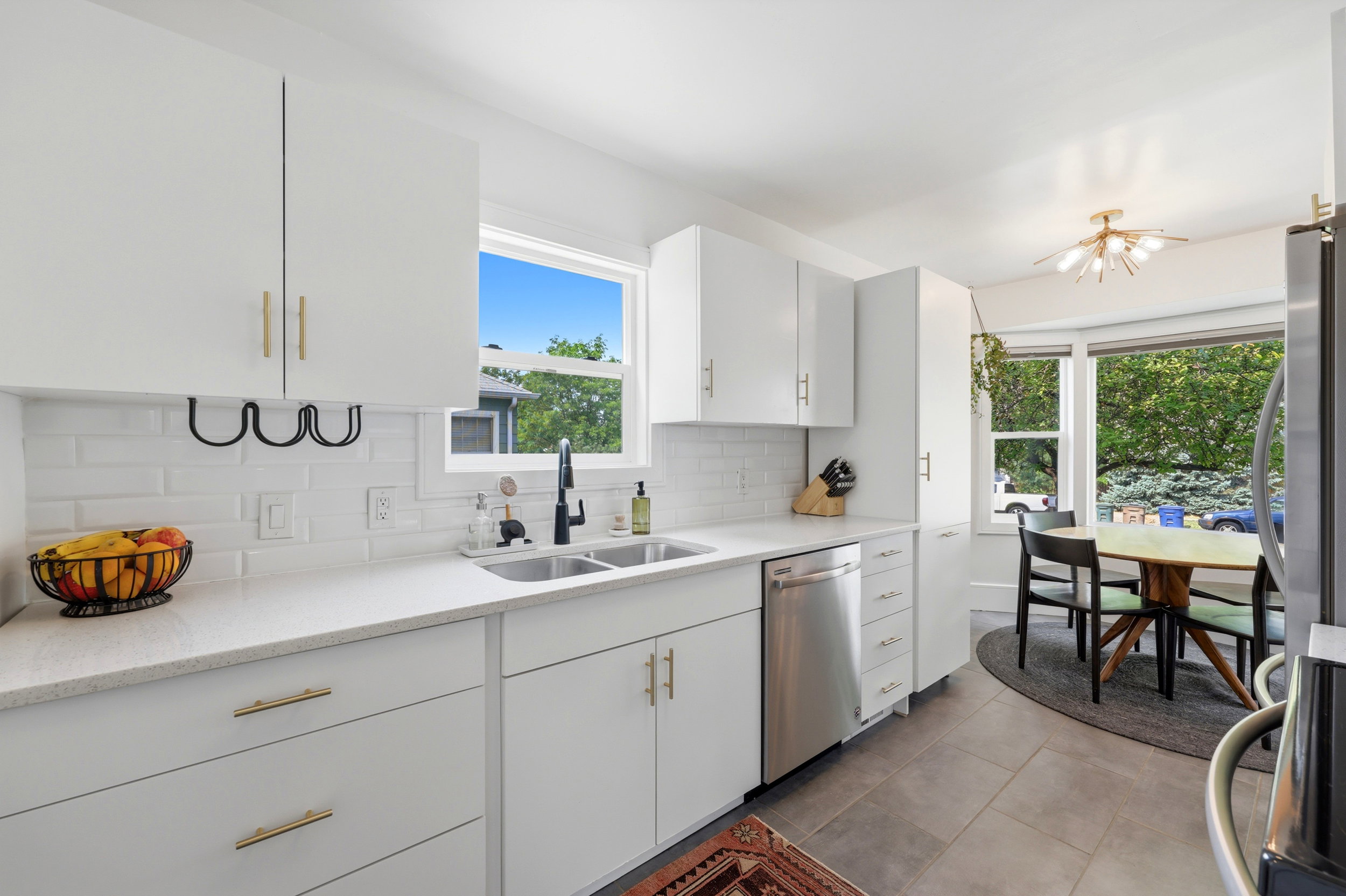 Bright kitchen featuring white cabinetry with gold-tone hardware, white subway tile backsplash, stainless steel double basin sink with matte black faucet, and durable tile flooring
