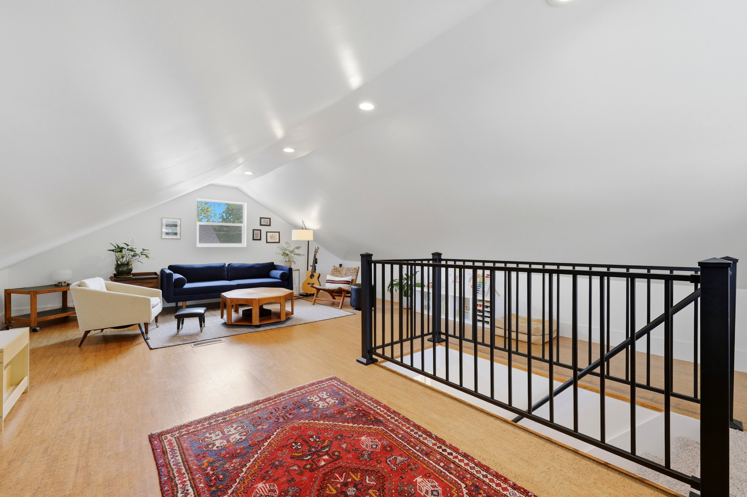 Spacious upper-level area featuring light wood-finish flooring, a contemporary black metal railing, a window providing natural light, recessed ceiling lighting, and sloped ceilings