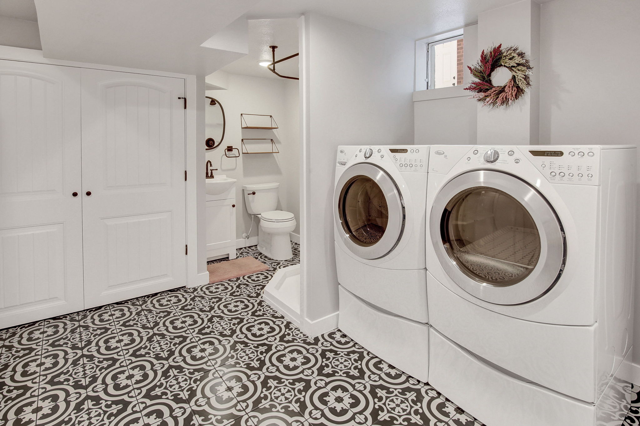 Laundry area featuring a patterned tile floor, white shaker-style cabinet doors, a pedestal sink with a matte black faucet, and a shower with a bronze-toned curtain rod