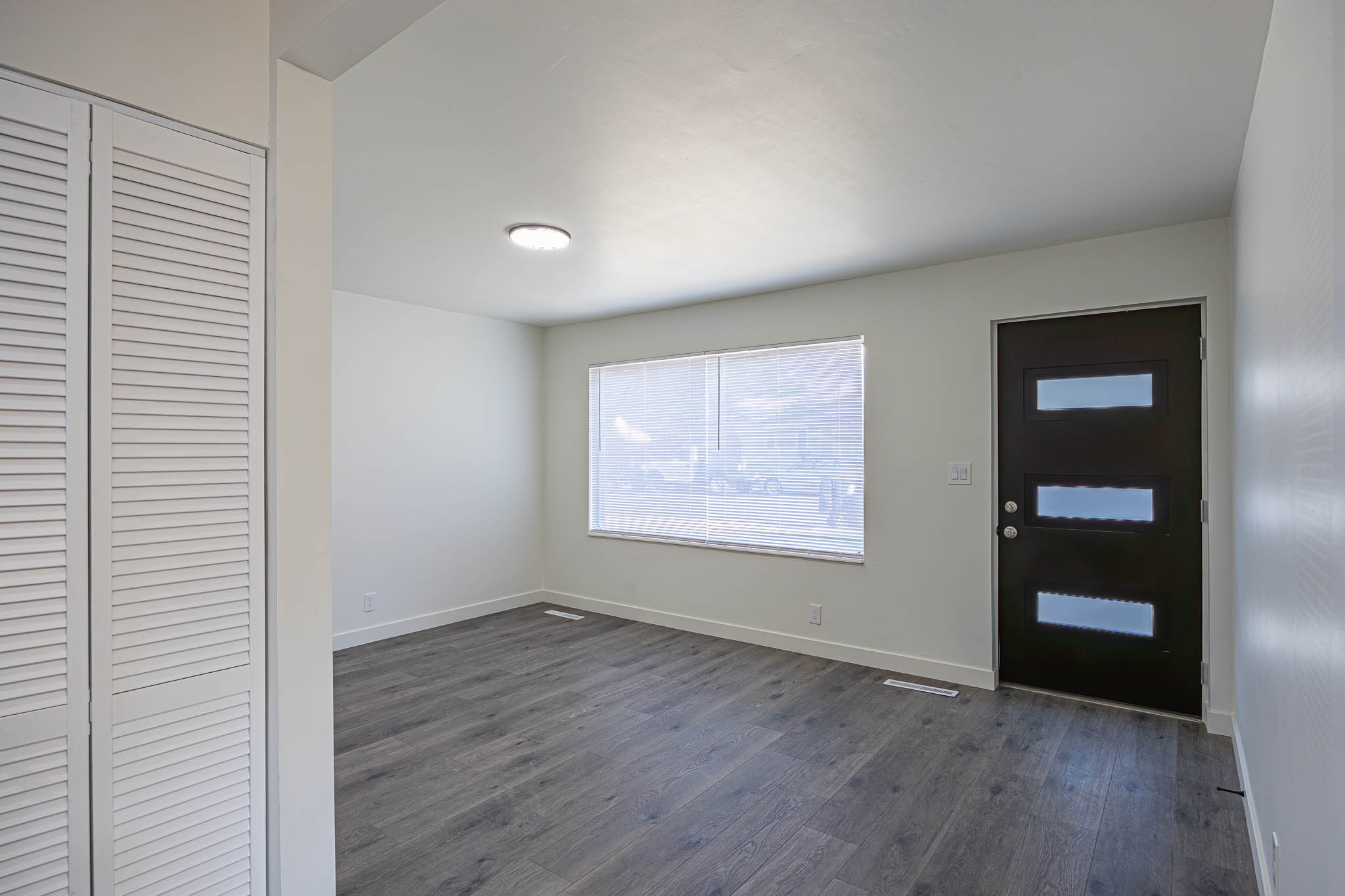 Entry area featuring wood-finish flooring, a contemporary dark entry door with frosted glass inserts, a window with horizontal blinds, a flush-mount ceiling light fixture, and white louvered bifold closet doors