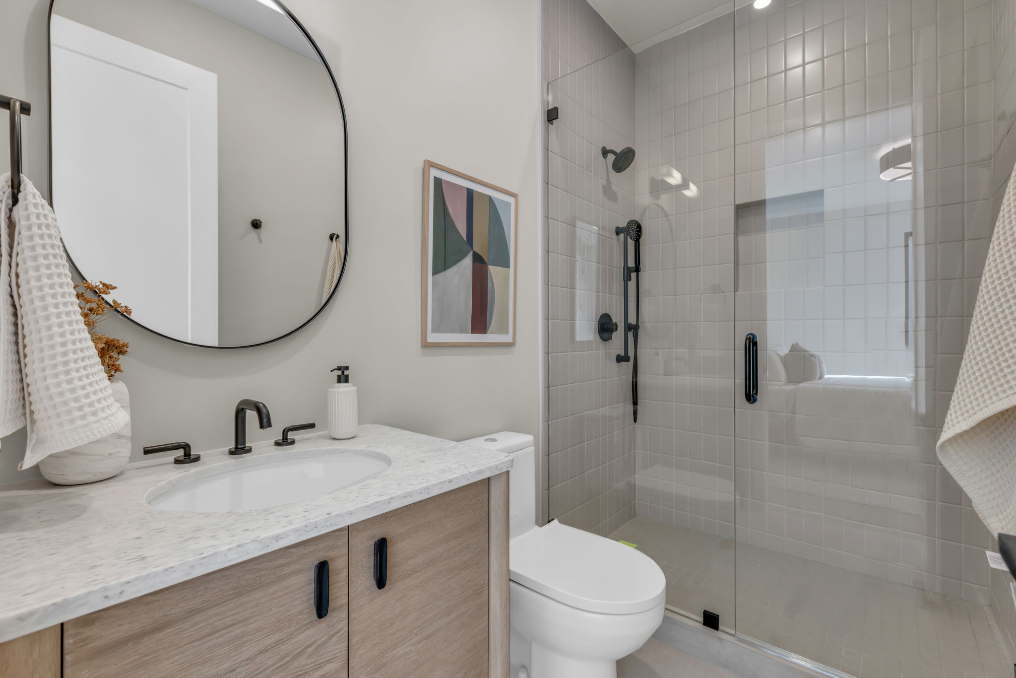 Bathroom featuring a wood-finish vanity with a light-toned countertop, an oval-shaped mirror, and a glass-enclosed shower with subway tile surround and matte black fixtures