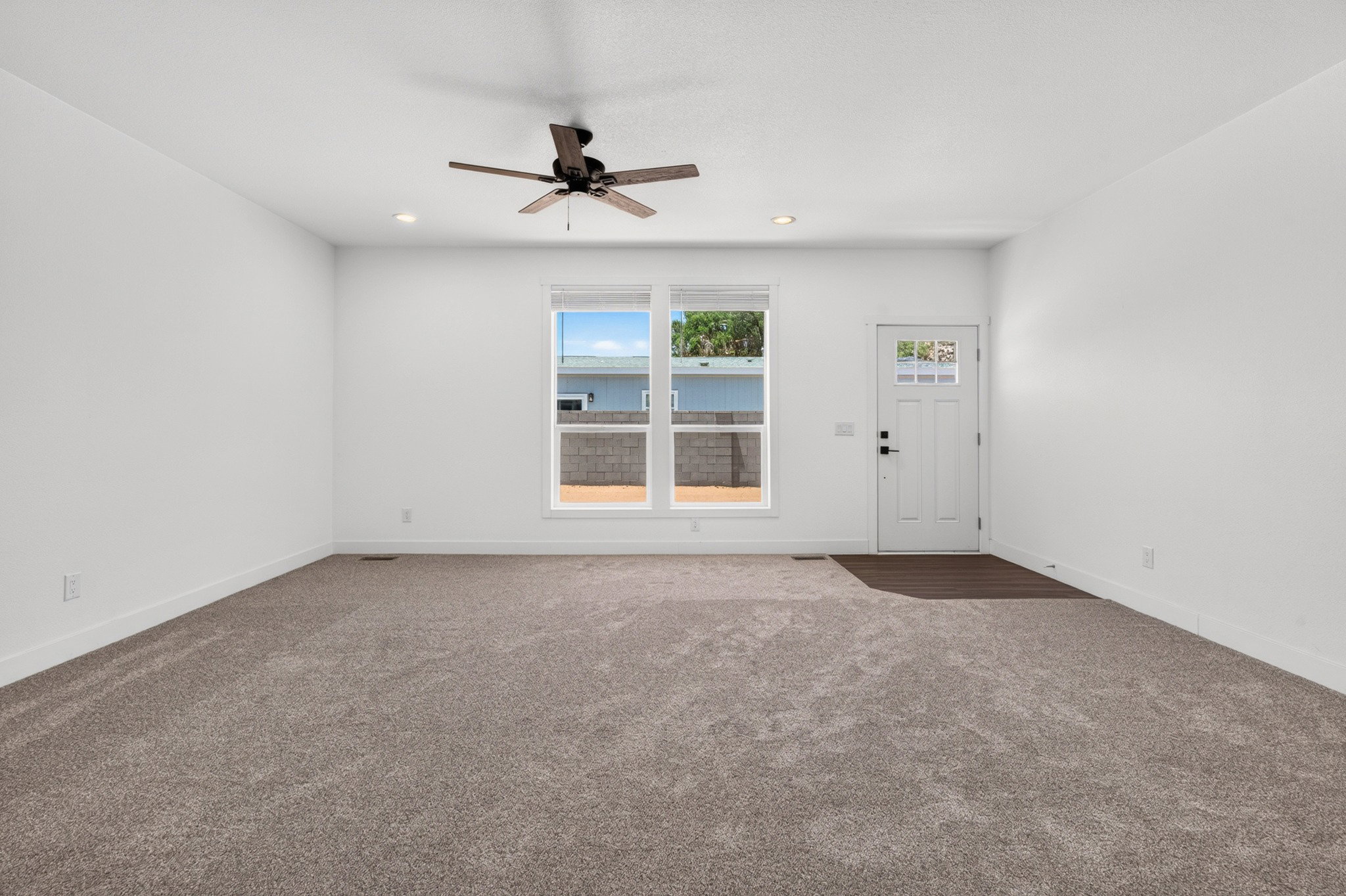 Spacious room featuring neutral carpeting, white walls, a multi-pane front door, a large window, and a ceiling fan with integrated lighting