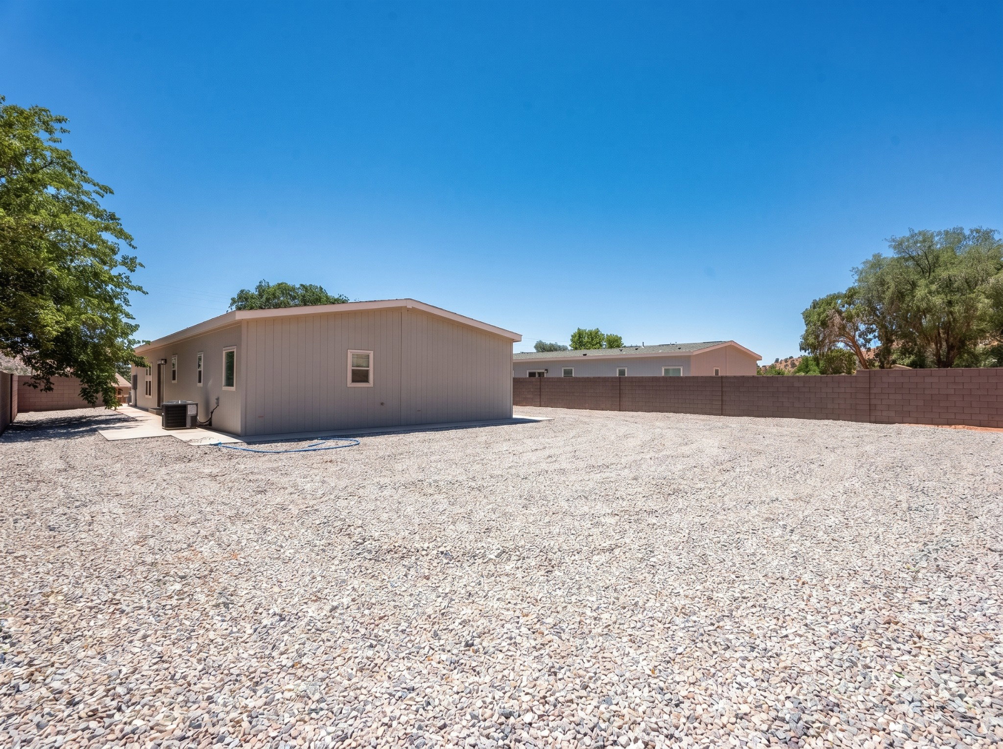 Extensive gravel yard offering a spacious footprint, featuring a light-colored stucco exterior with vertical siding accents, multiple windows, and a concrete patio area