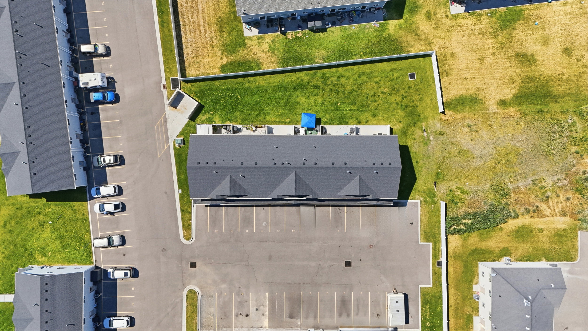 Multi-unit residential structure featuring a dark shingle roof, integrated parking lot with marked spaces, surrounding green spaces, paved driveways, and a nearby row of similar buildings