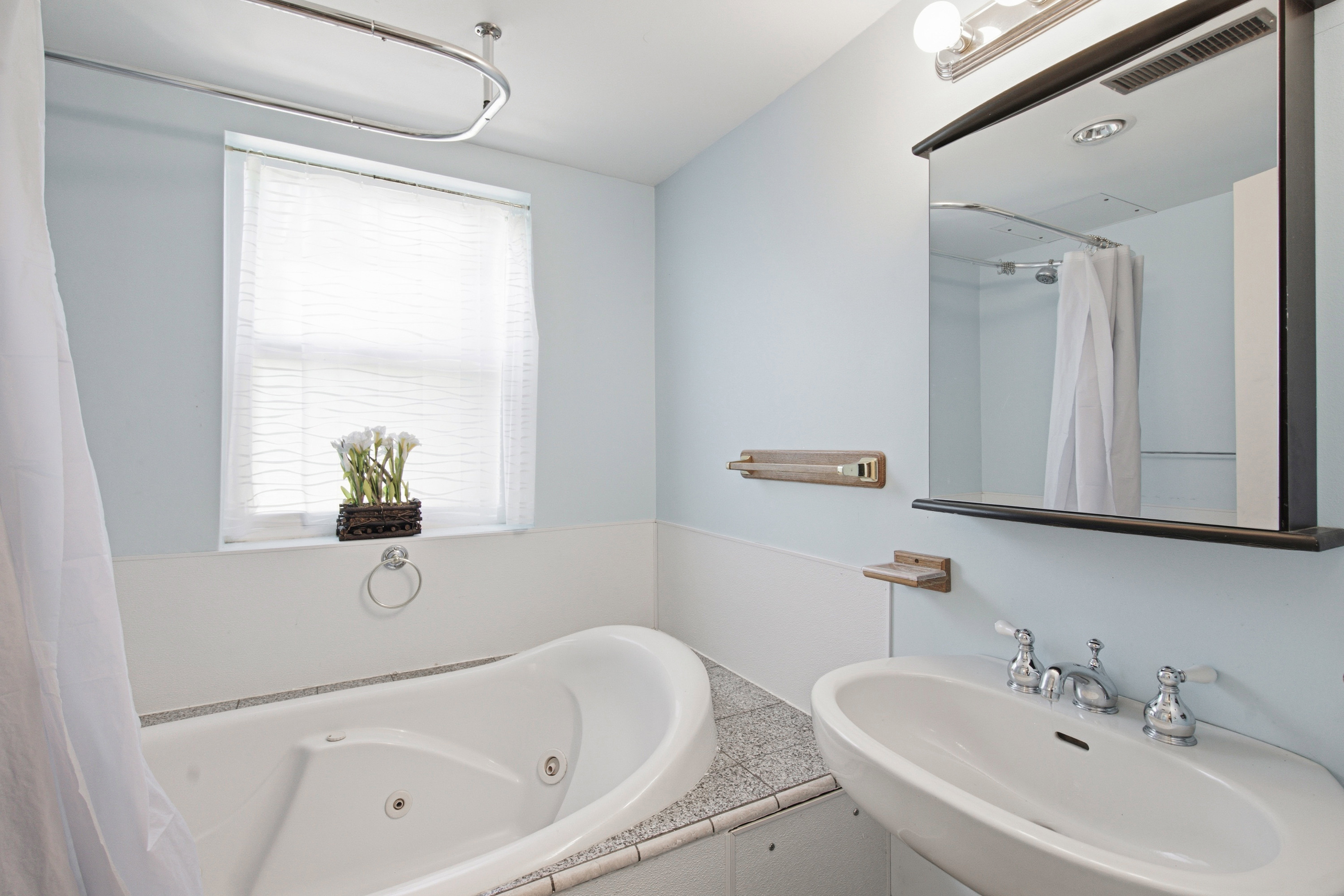 Bathroom featuring a jetted tub, a window with blinds, a wall-mounted sink with chrome fixtures, a framed mirror, and a wall-mounted light fixture