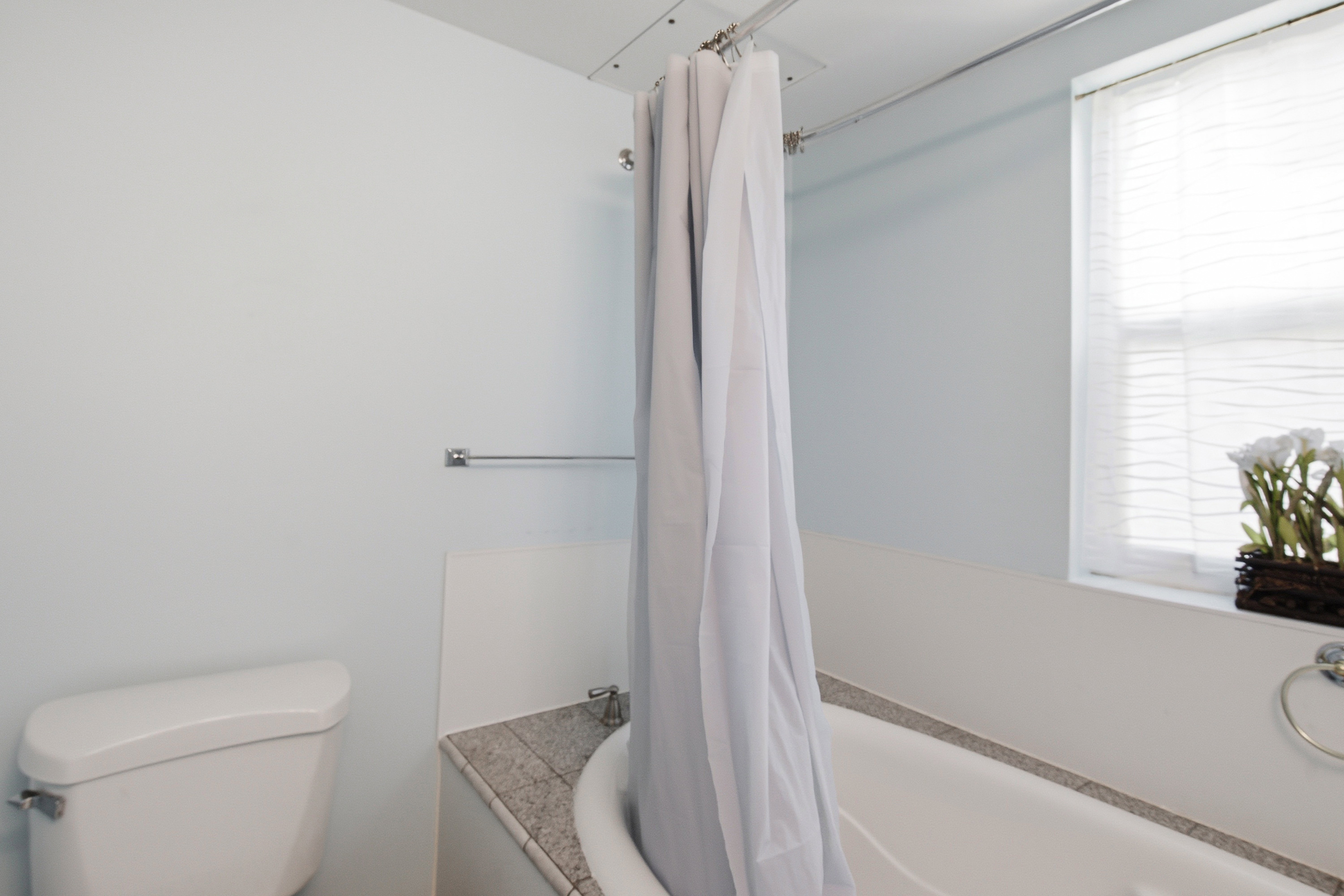 Bathroom featuring a white porcelain toilet, a white bathtub with a stone-finish surround, a window with horizontal blinds, and a chrome towel ring