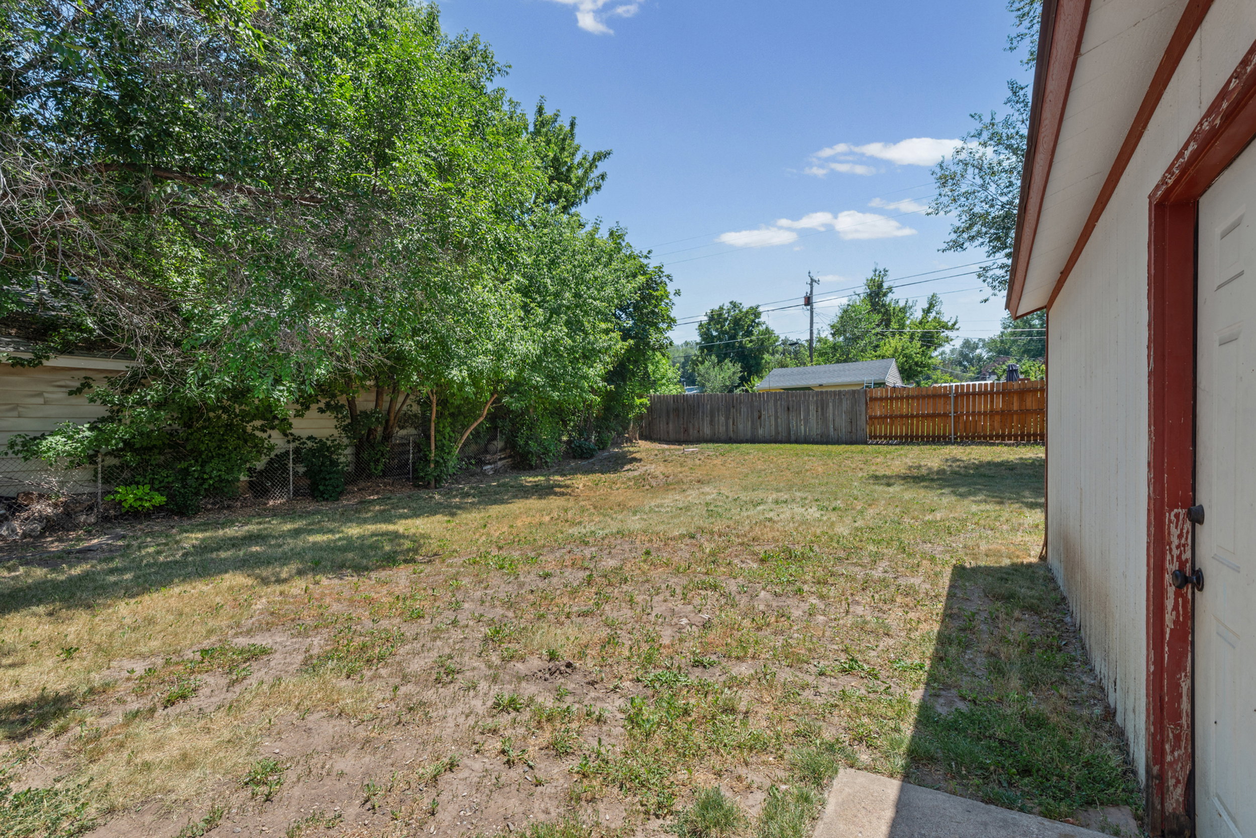 Expansive backyard featuring a combination wood and chain-link perimeter fence, mature trees and shrubs, and a detached outbuilding with a white exterior and red trim