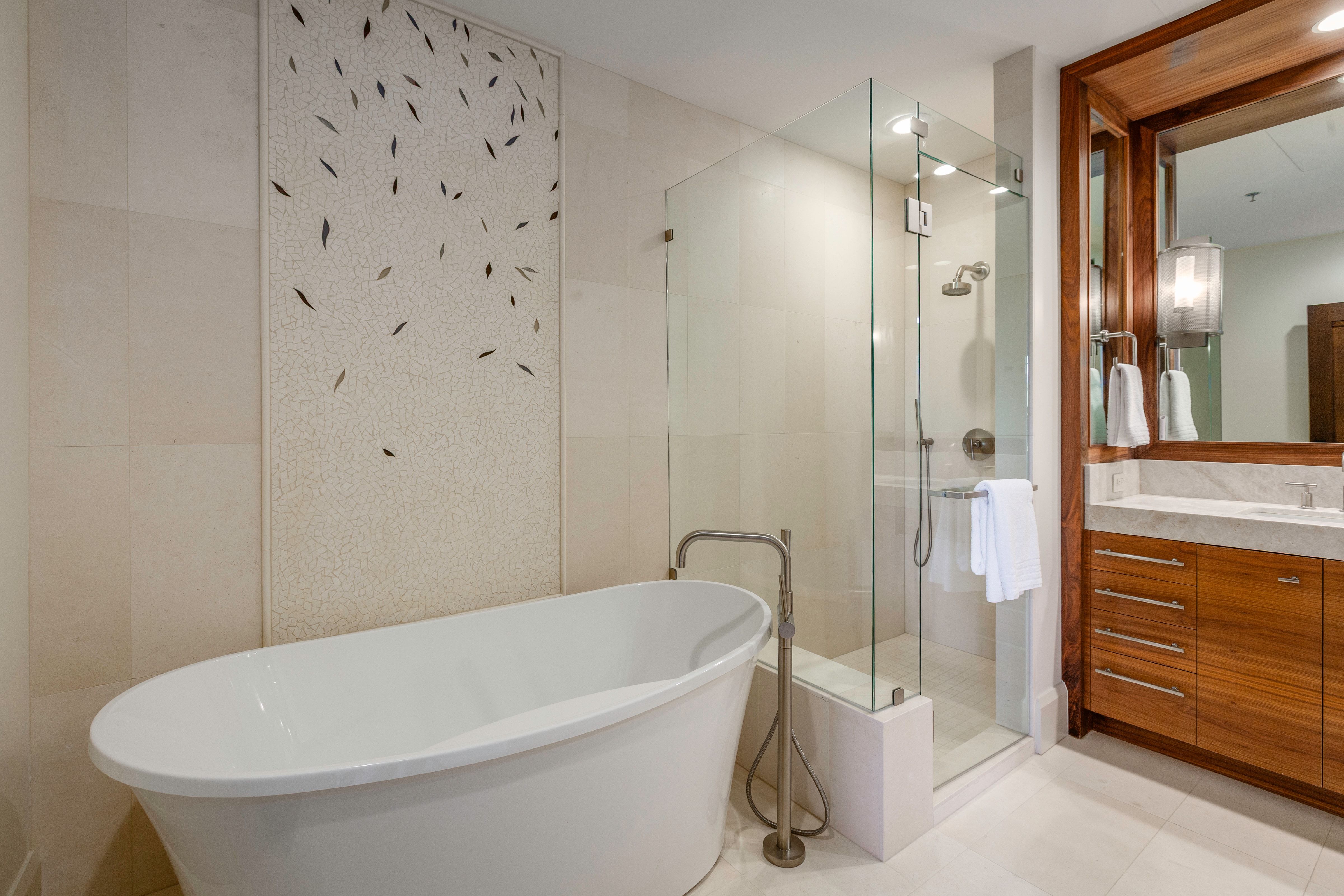 Freestanding soaking tub with floor-mounted faucet, glass-enclosed shower, wood-finish vanity with stone countertop, large mirror, and tiled accent wall