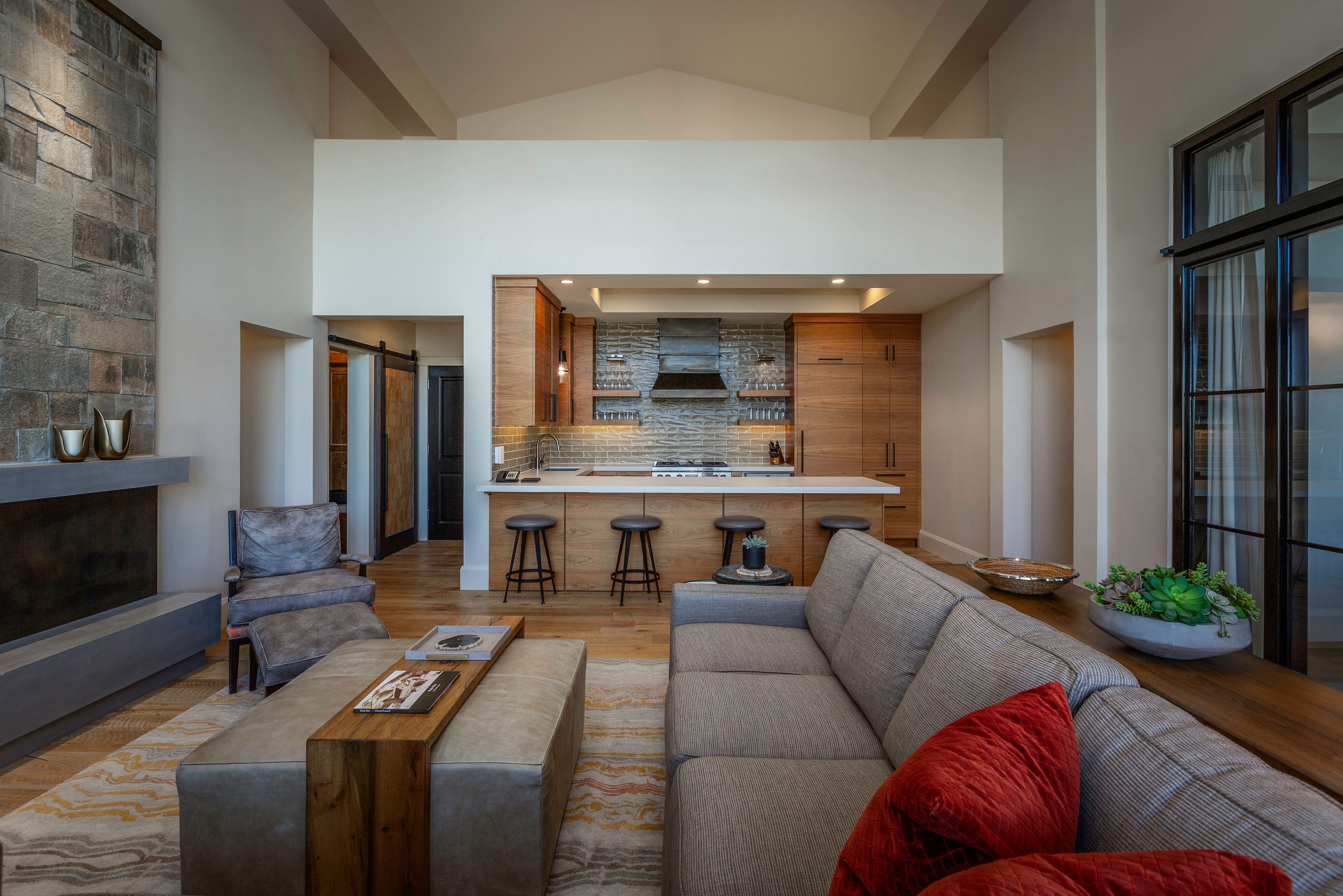 Open-concept living space featuring a floor-to-ceiling stone fireplace, a kitchen with wood cabinetry and a white countertop, and high vaulted ceilings with exposed beams