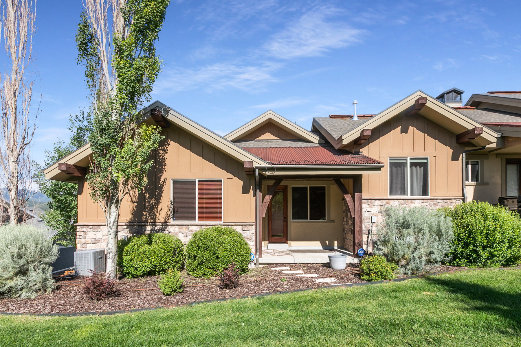 Mountain-style exterior featuring a stone and board and batten facade, covered entry with exposed wood beams, red corrugated metal accent roof, and a manicured lawn with mature landscaping