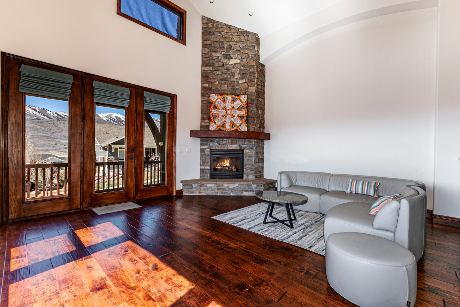 Spacious living area featuring rich wood-finish flooring, a stone fireplace with a substantial wood mantel, a vaulted ceiling, and multiple glass-paneled doors with direct outdoor access