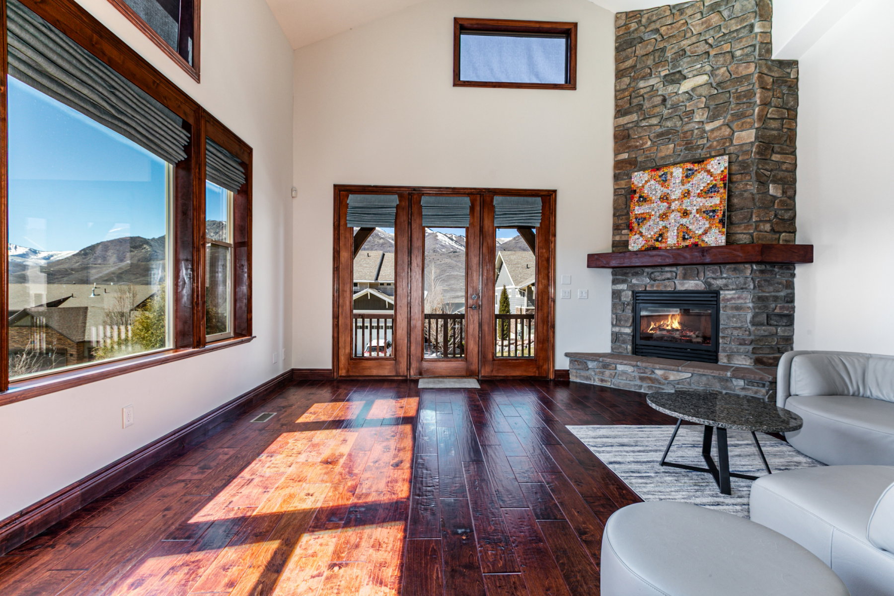 Spacious living area featuring a stone fireplace with a substantial wood mantle, vaulted ceilings, expansive windows with mountain scenery, and dark wood-finish flooring