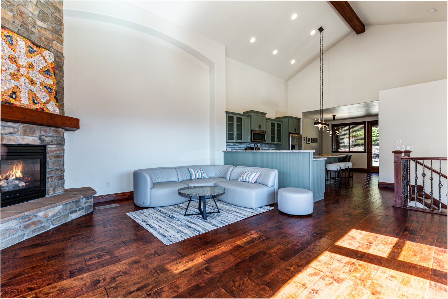 Spacious living area featuring rich wood-finish flooring, a stone fireplace with a substantial wood mantel, vaulted ceilings with exposed wood beams, and recessed lighting