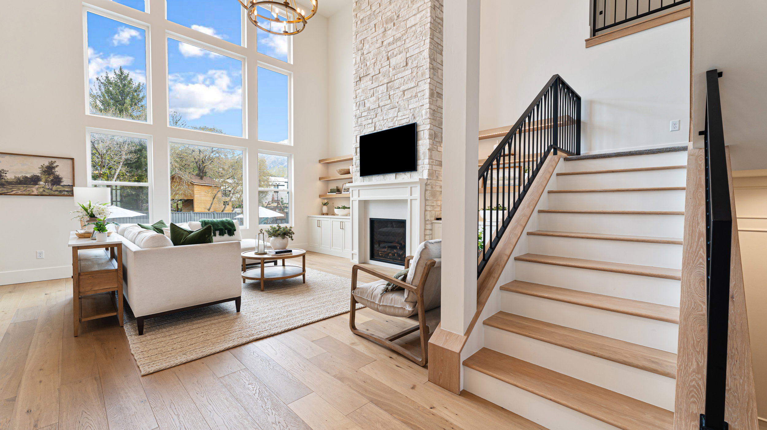 Two-story living area featuring extensive window walls, wood-finish flooring, a stacked stone fireplace with a white mantel, built-in shelving, and a prominent staircase with black metal railings