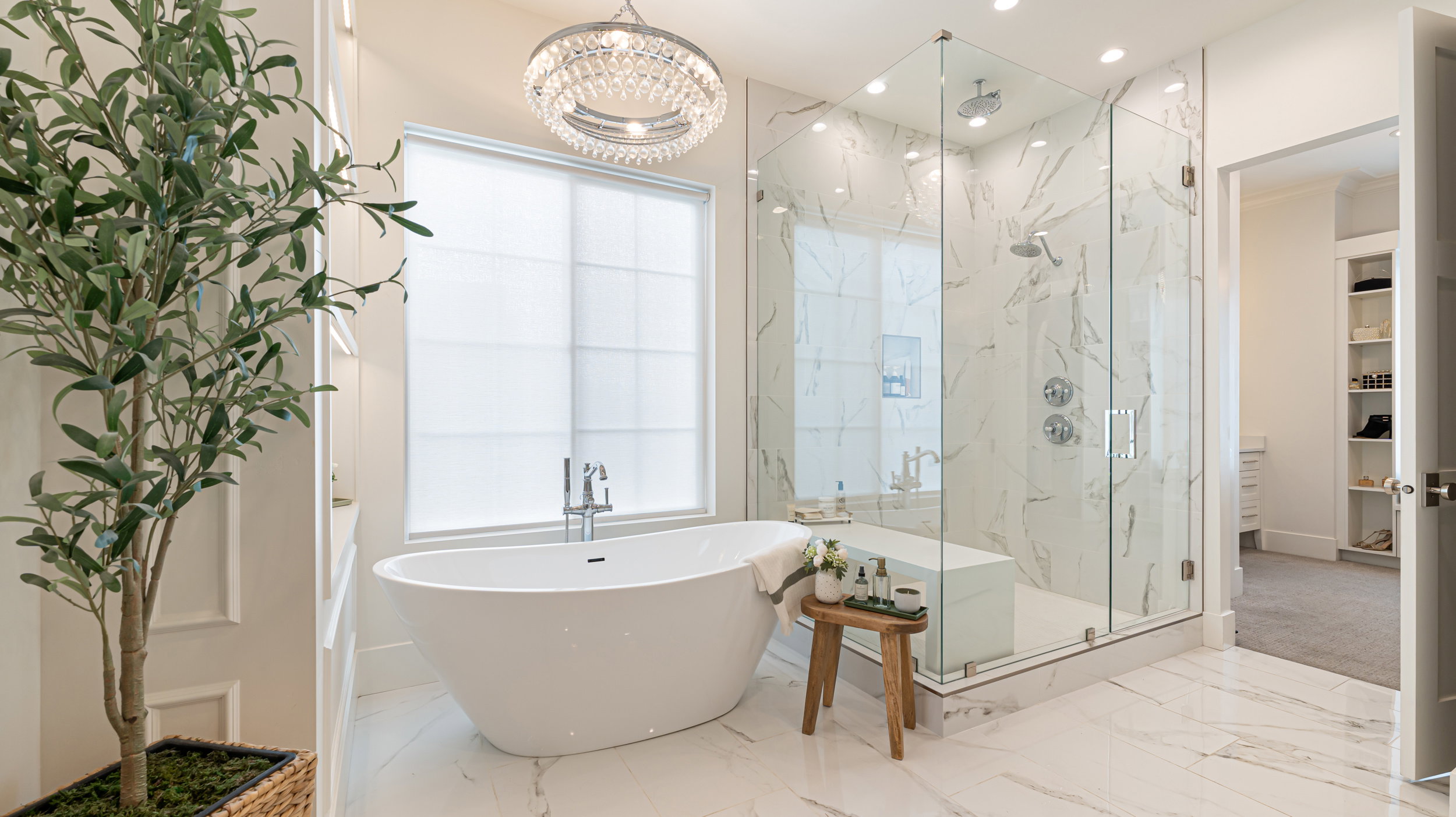Luxurious bathroom featuring a freestanding soaking tub, a spacious glass-enclosed shower with multiple showerheads and a built-in bench, marble-finish wall and floor tiling, and a crystal chandelier