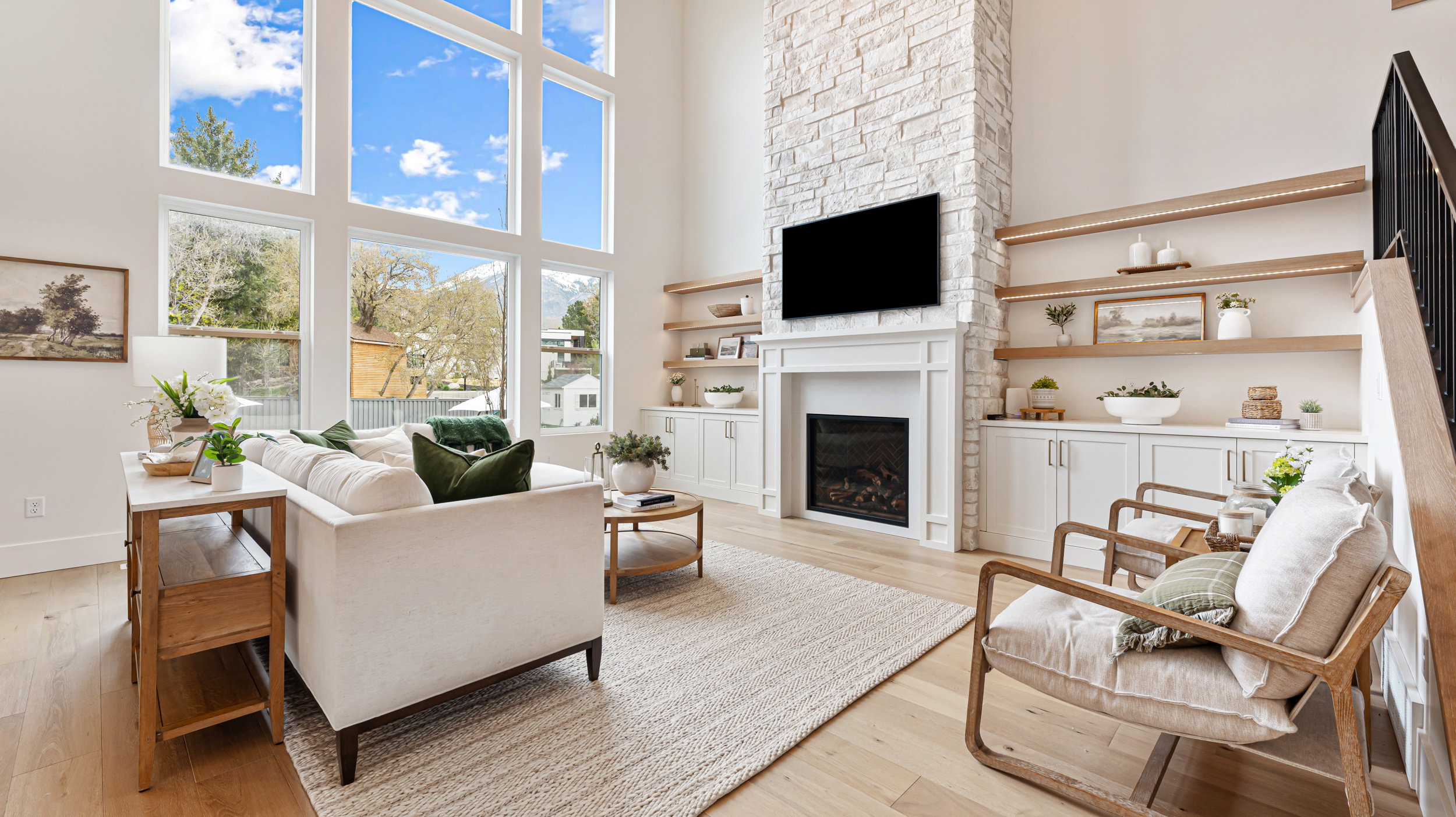 Two-story living area featuring a stacked stone fireplace with a white mantel, custom built-in cabinetry, wood-finish flooring, and a dramatic floor-to-ceiling window wall