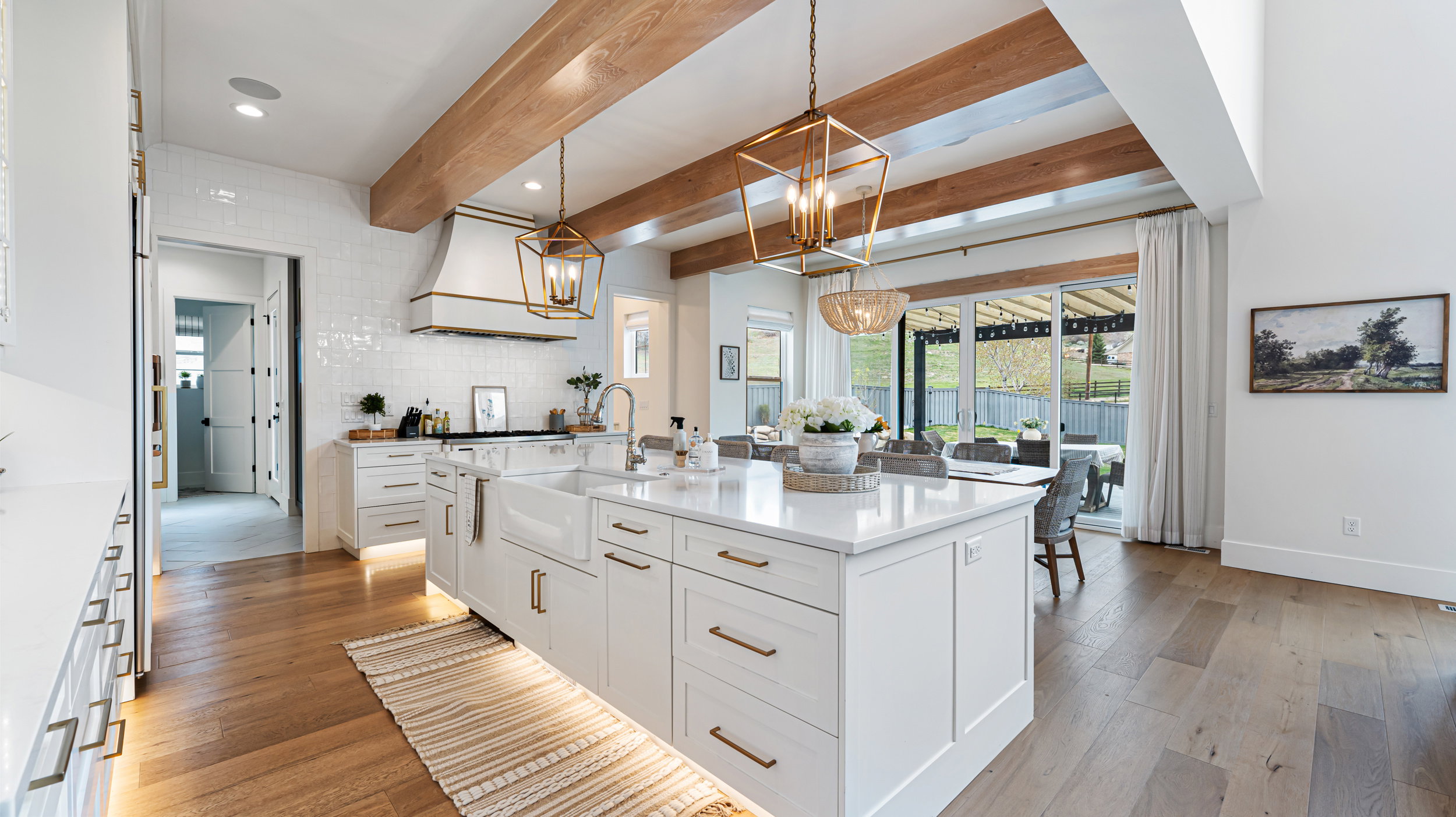 Gourmet kitchen featuring a large central island with an integrated farmhouse sink, white cabinetry with brass hardware, wood-finish flooring, and exposed wood ceiling beams
