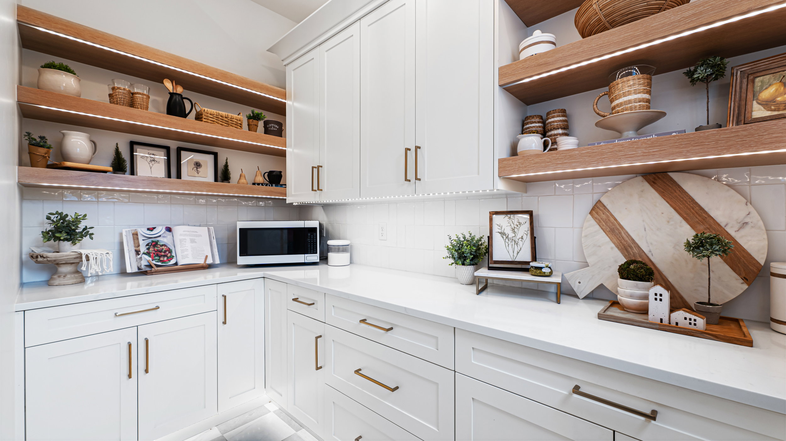 White cabinetry with gold-tone hardware, white subway tile backsplash, white countertops, and integrated wood shelving with accent lighting