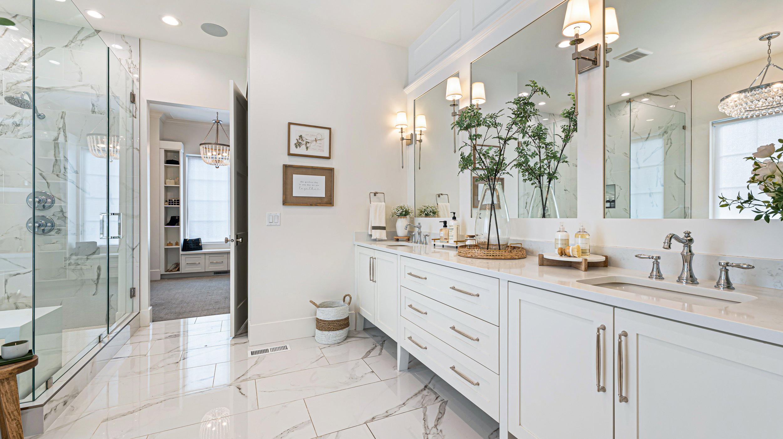 Spacious bathroom featuring a dual vanity with white cabinetry and undermount sinks, a glass-enclosed shower with a bench and subway tile surround, large format tile flooring, and multiple wall sconces with mirrored accents