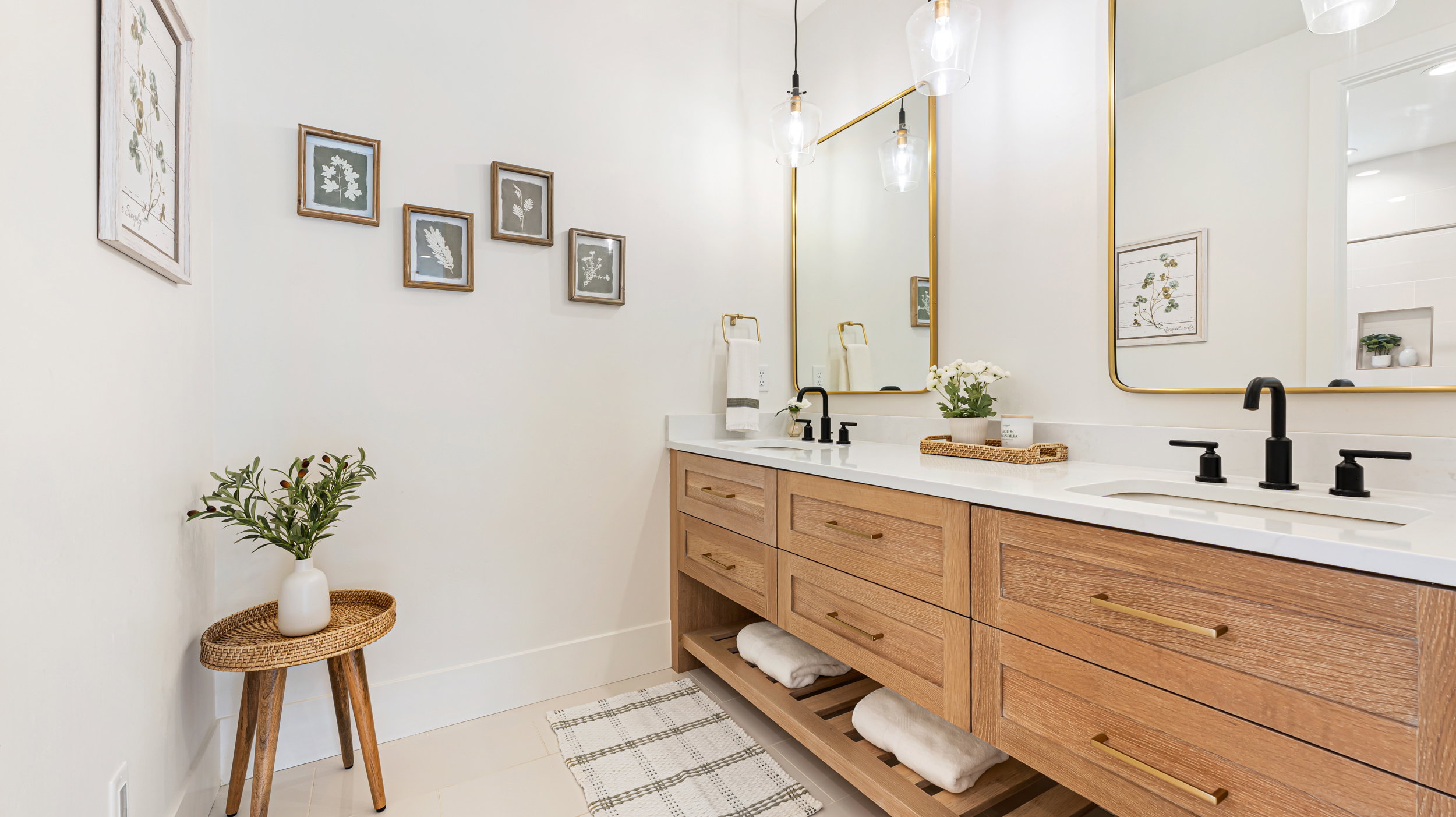 Dual vanity with light wood-finish cabinetry, white countertops, matte black fixtures, and gold-framed mirrors