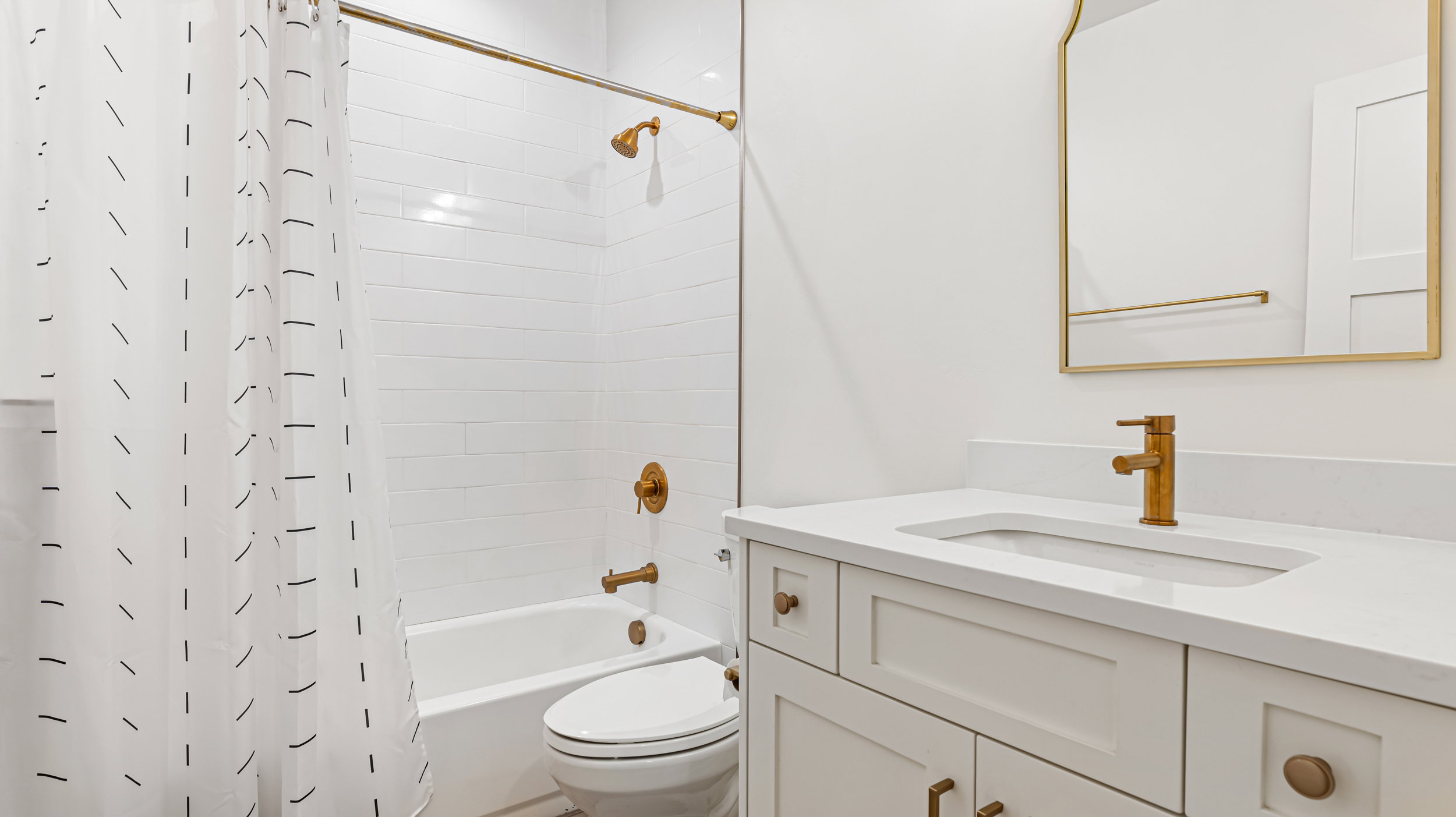Bathroom featuring a white subway tile shower surround, brass-finish fixtures, white vanity with recessed panel doors, integrated sink, and a white countertop