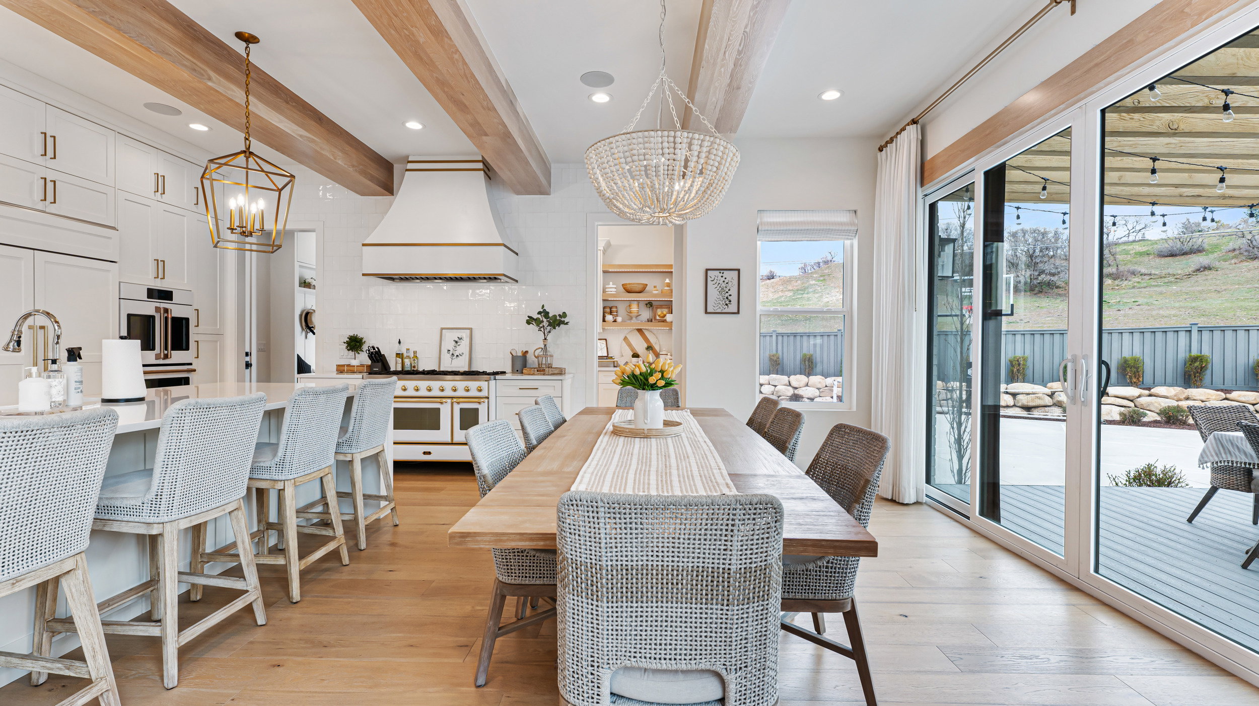 Open-concept kitchen and dining area featuring light wood-finish flooring, exposed wood ceiling beams, a large kitchen island with seating, white cabinetry, and a sliding glass door opening to an outdoor deck