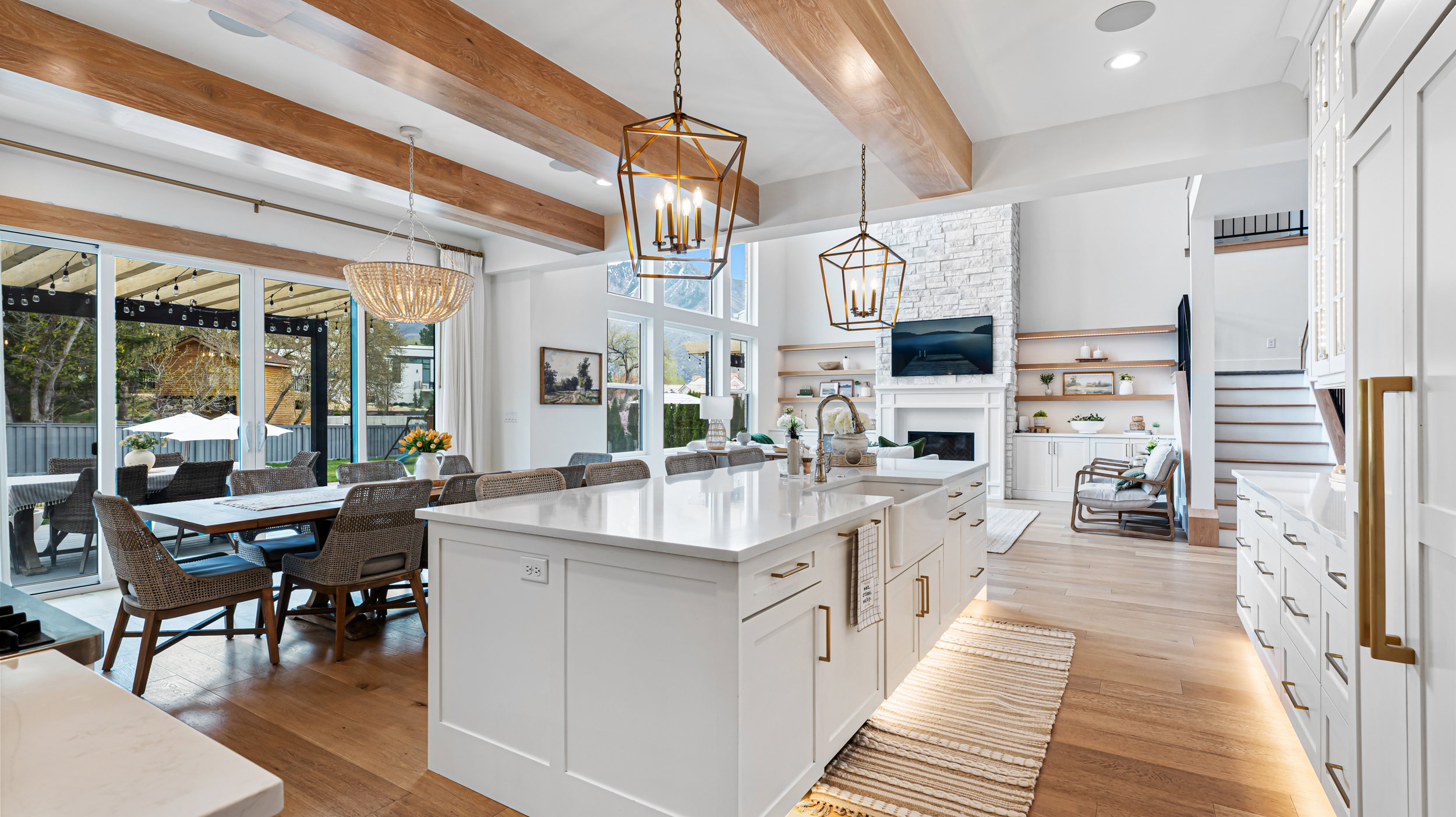 Open-concept interior featuring light wood-finish flooring, exposed ceiling beams, white cabinetry with gold-tone hardware, a large kitchen island with a farmhouse sink, and sliding glass doors leading to an outdoor dining area