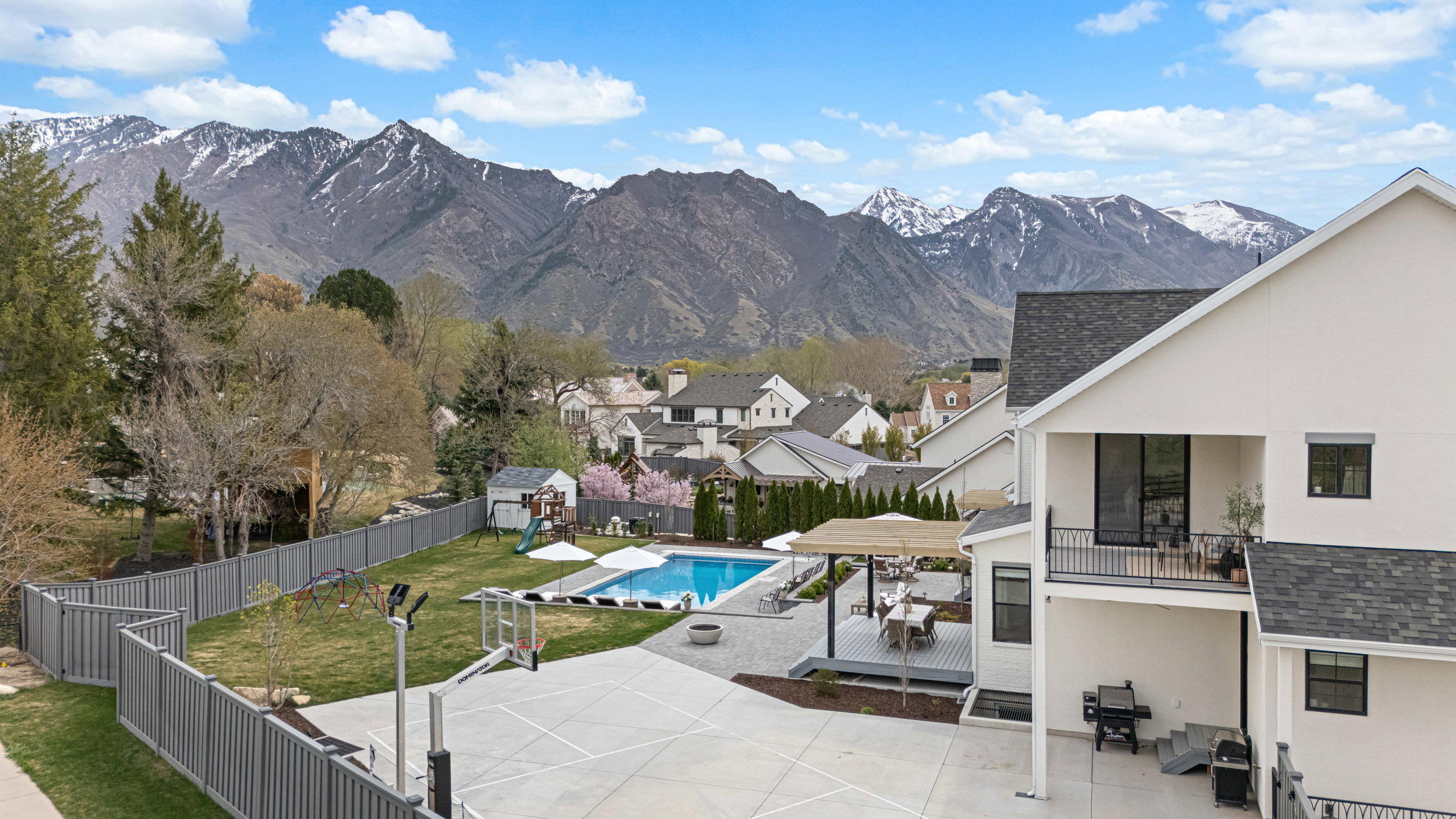 Expansive backyard featuring a swimming pool, adjacent paved patio with a pergola, a dedicated sports court, and a grassy lawn, all framed by a gray privacy fence