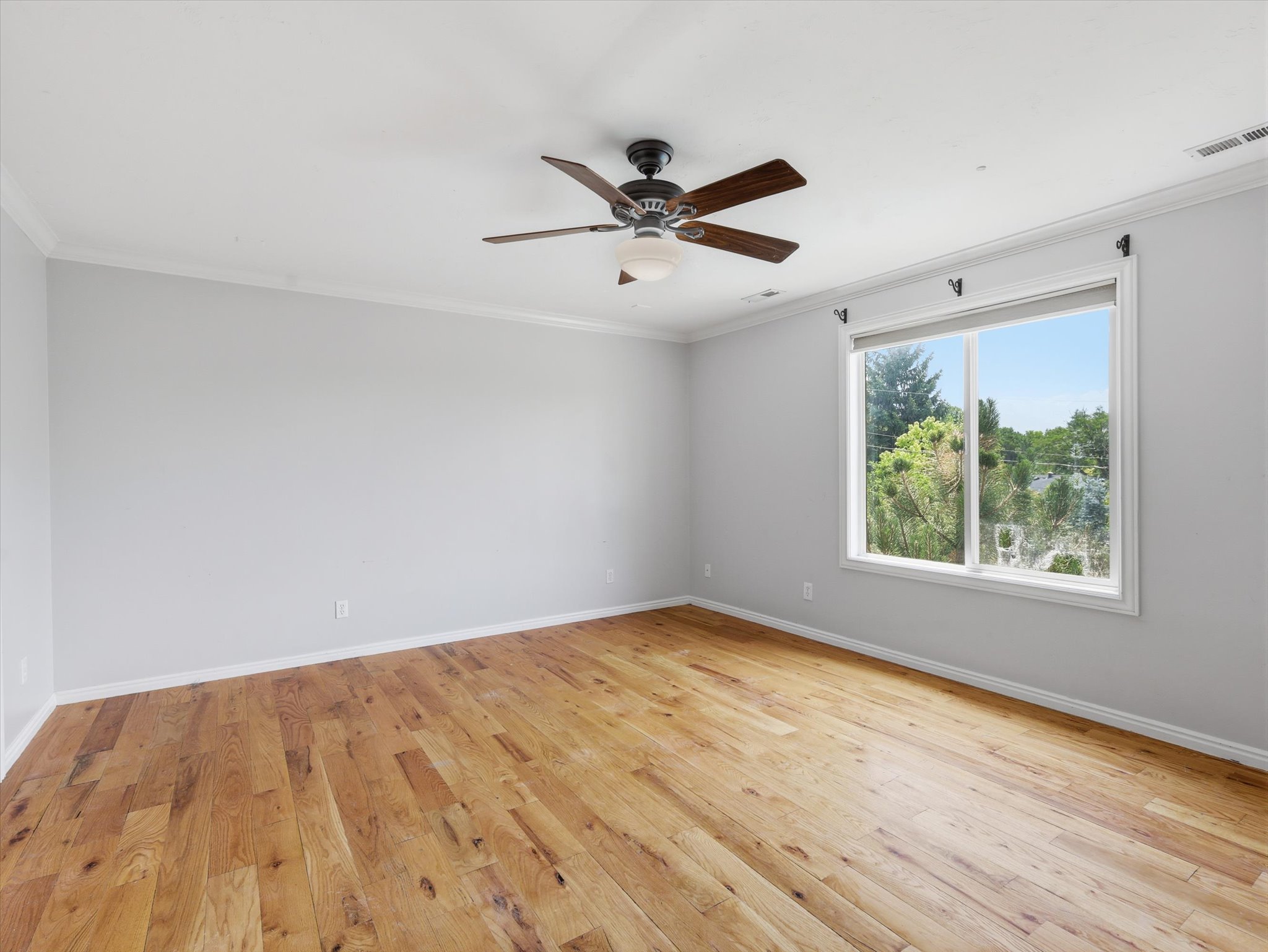Spacious room featuring wood-finish flooring, a large window, a ceiling fan with integrated lighting, crown molding, and neutral wall tones