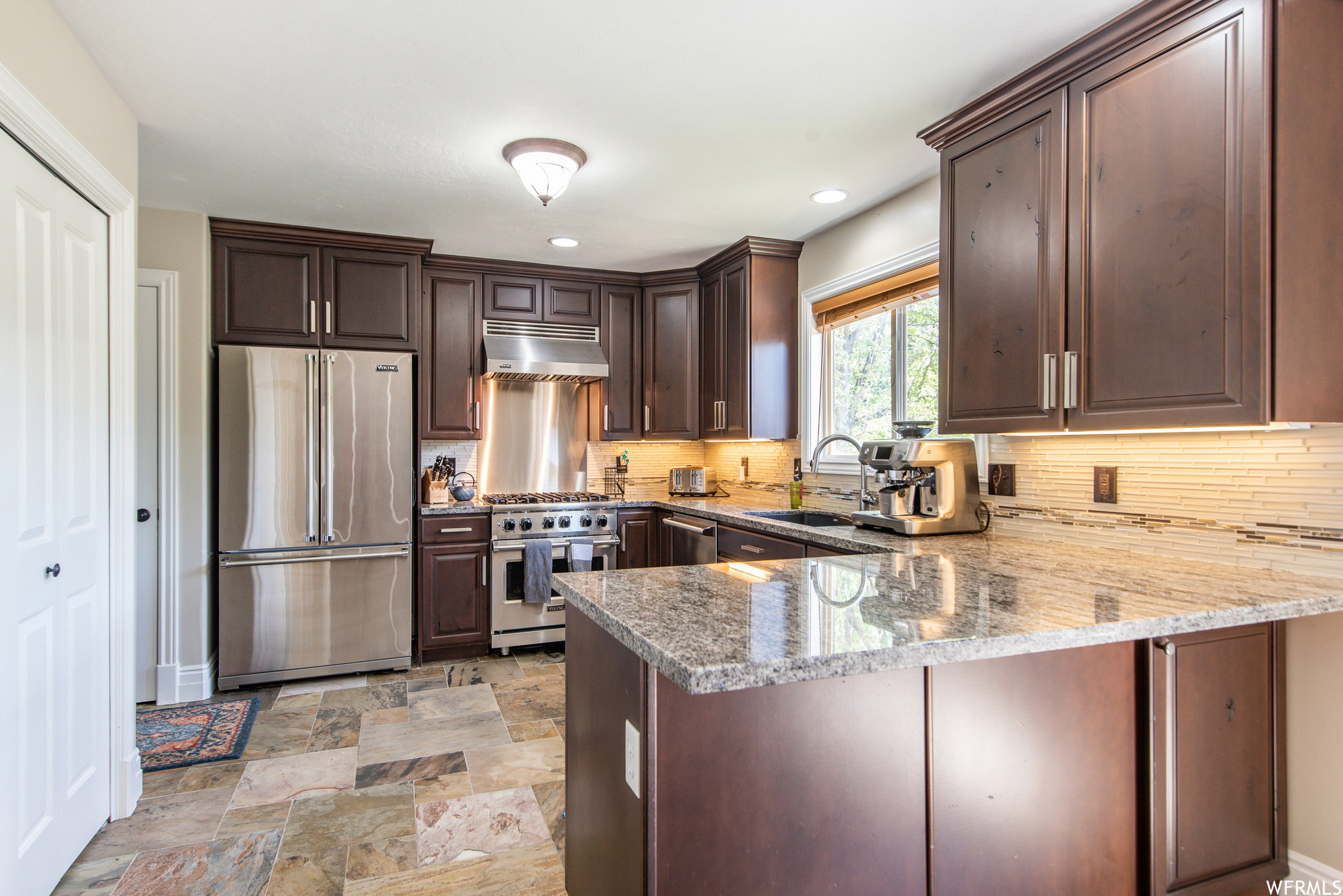 Kitchen featuring dark wood cabinetry, stainless steel appliances, speckled stone countertops, a tile backsplash, and multi-toned stone tile flooring