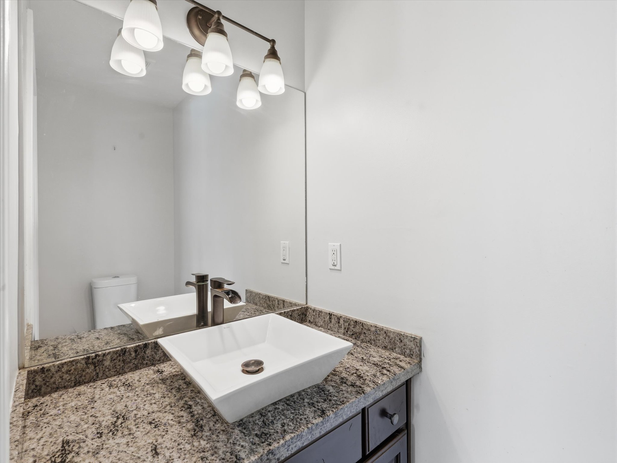Bathroom vanity featuring a rectangular vessel sink, granite-style countertop, dark wood-finish cabinetry, bronze-finish faucet, and coordinating bronze-finish light fixture with five frosted glass shades