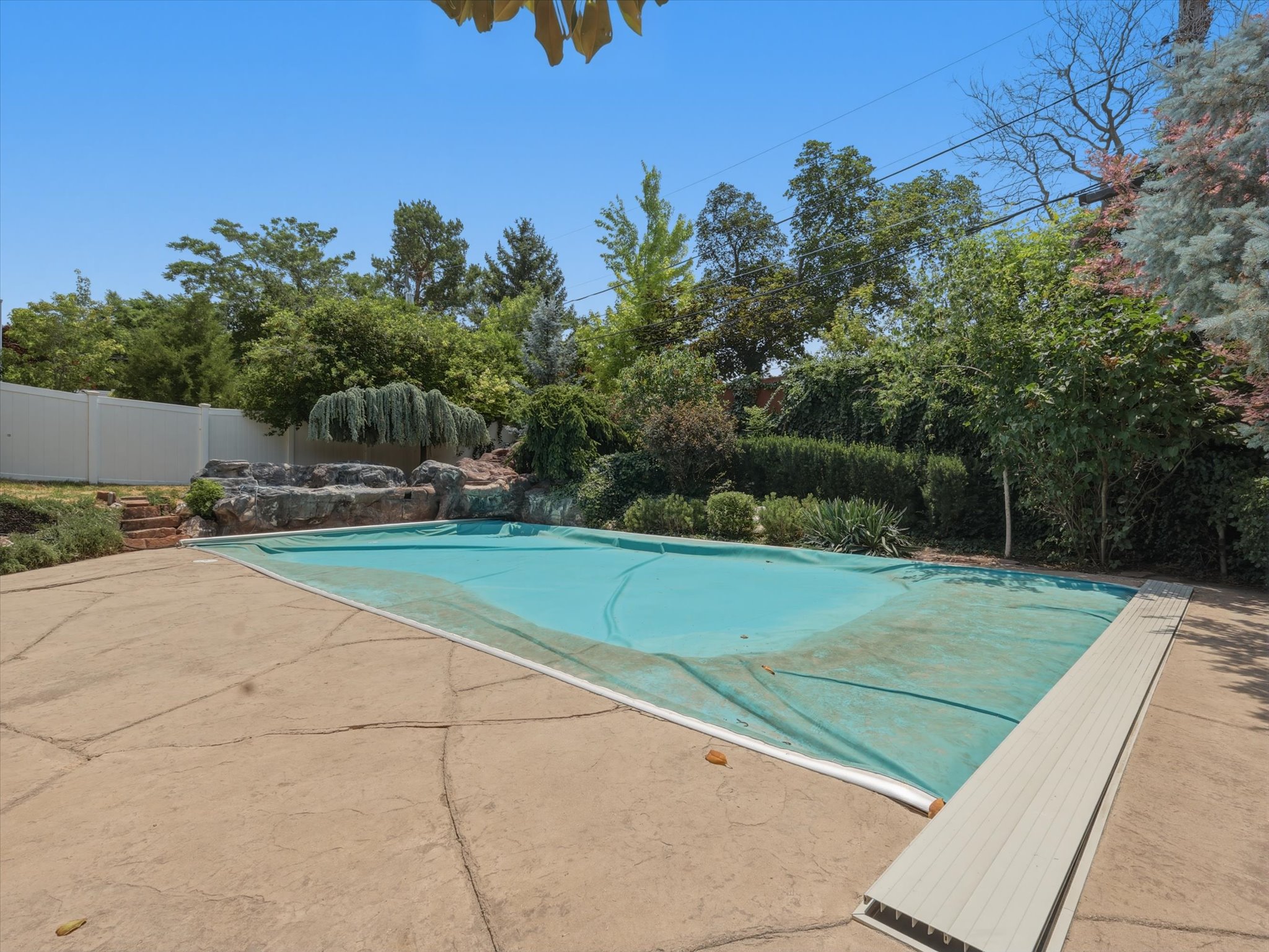 Outdoor swimming pool with a rock waterfall feature, surrounded by mature landscaping and a white privacy fence